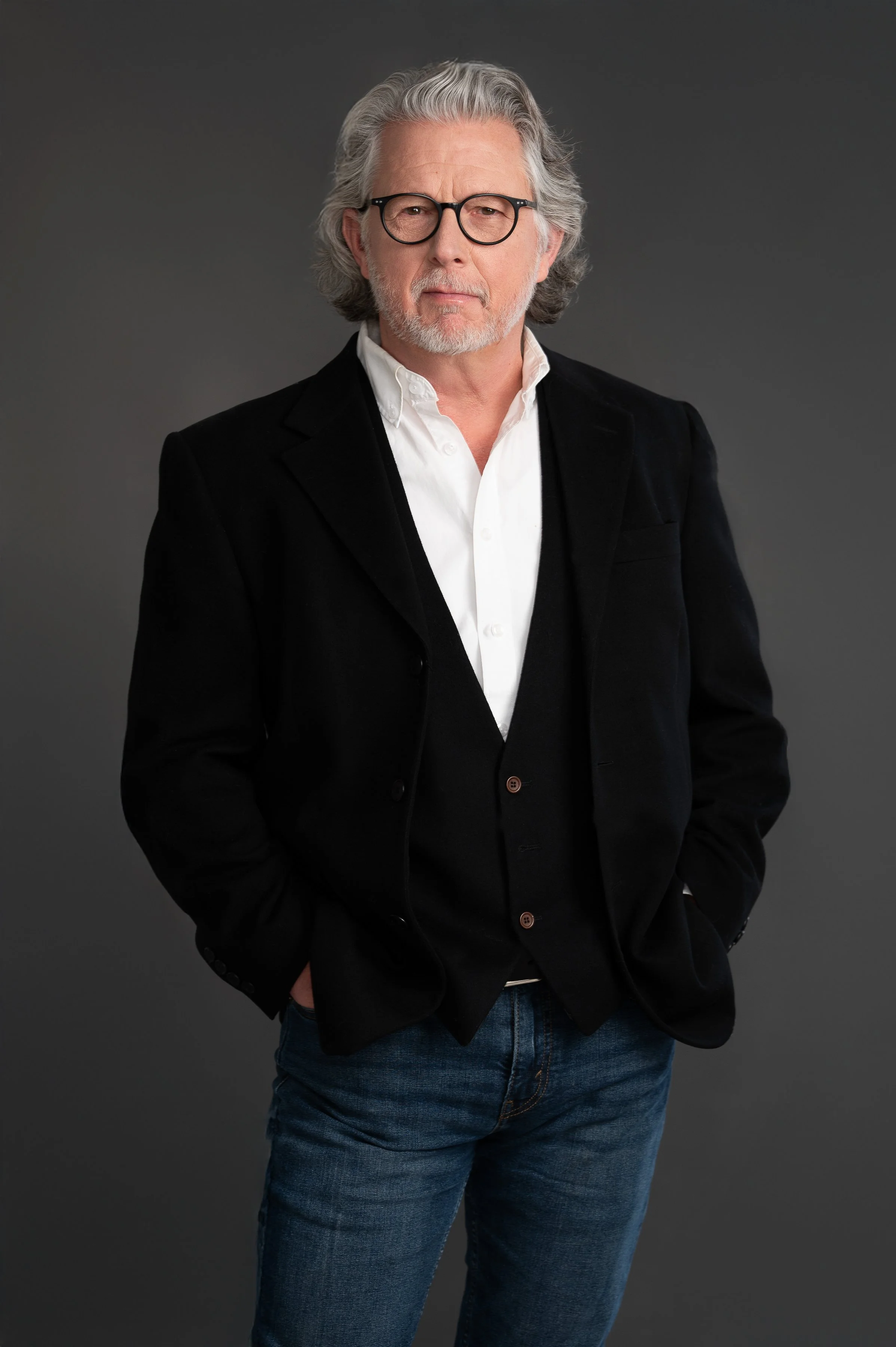 A man with gray hair, beard, and mustache wearing black-rimmed glasses, a white dress shirt, a black blazer, and blue jeans, standing against a gray background.