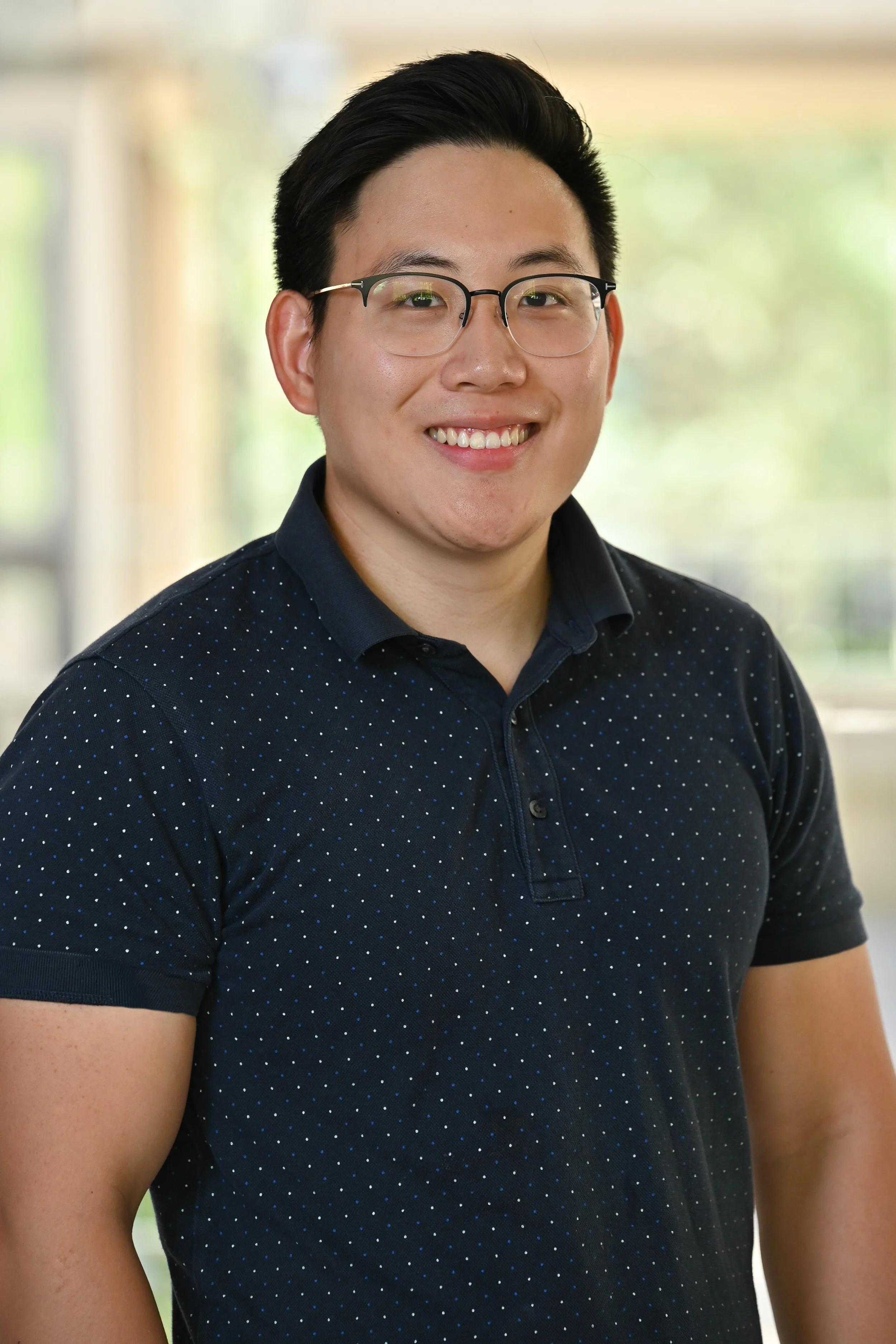 A young man with black hair and glasses smiling, wearing a navy blue polo shirt with small blue and white dots, standing indoors in front of a blurred background with green and beige tones.
