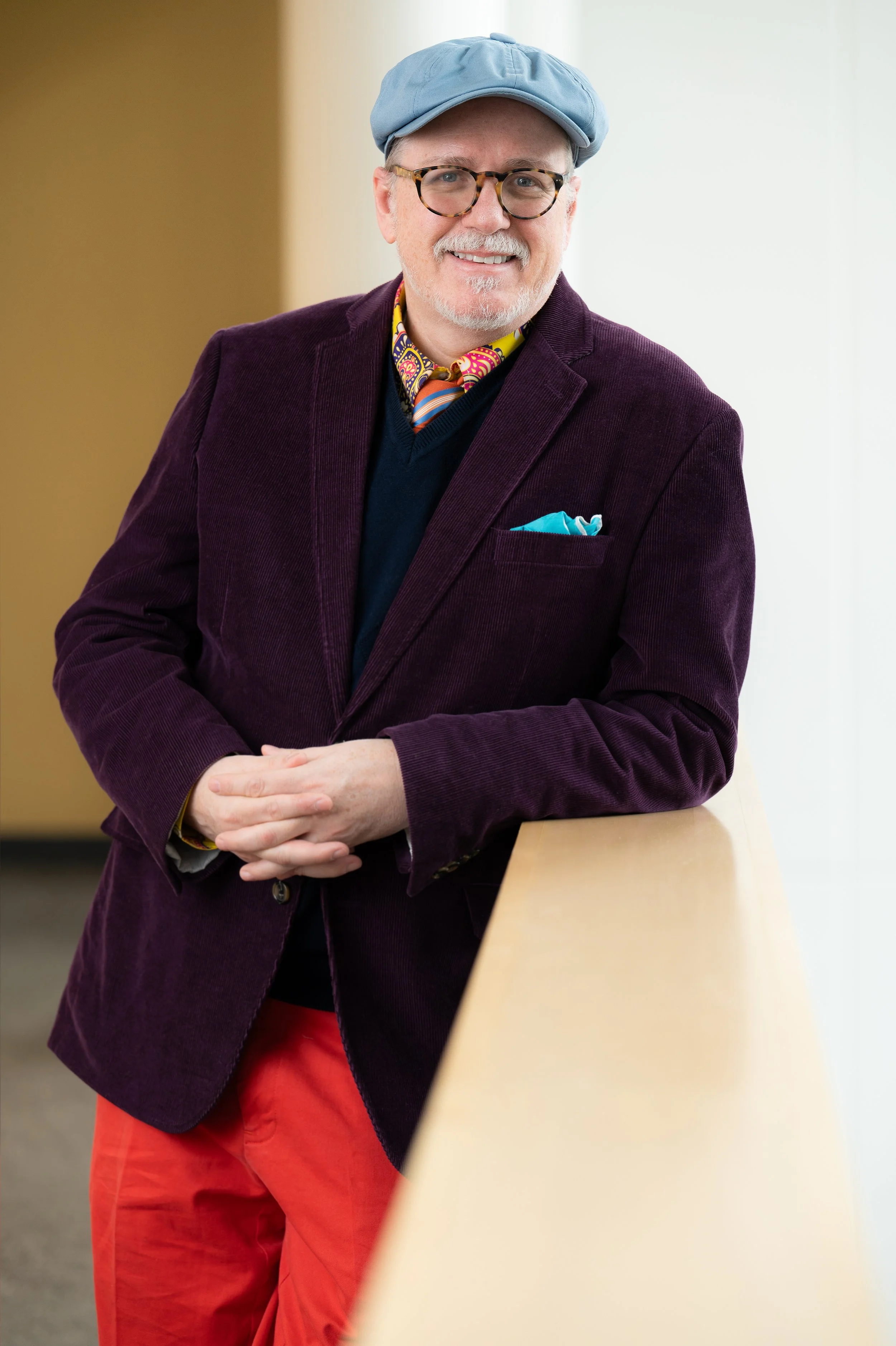 A man smiling, wearing glasses, a light blue cap, a patterned shirt, a velvet purple blazer, and red pants, standing beside a light-colored counter.