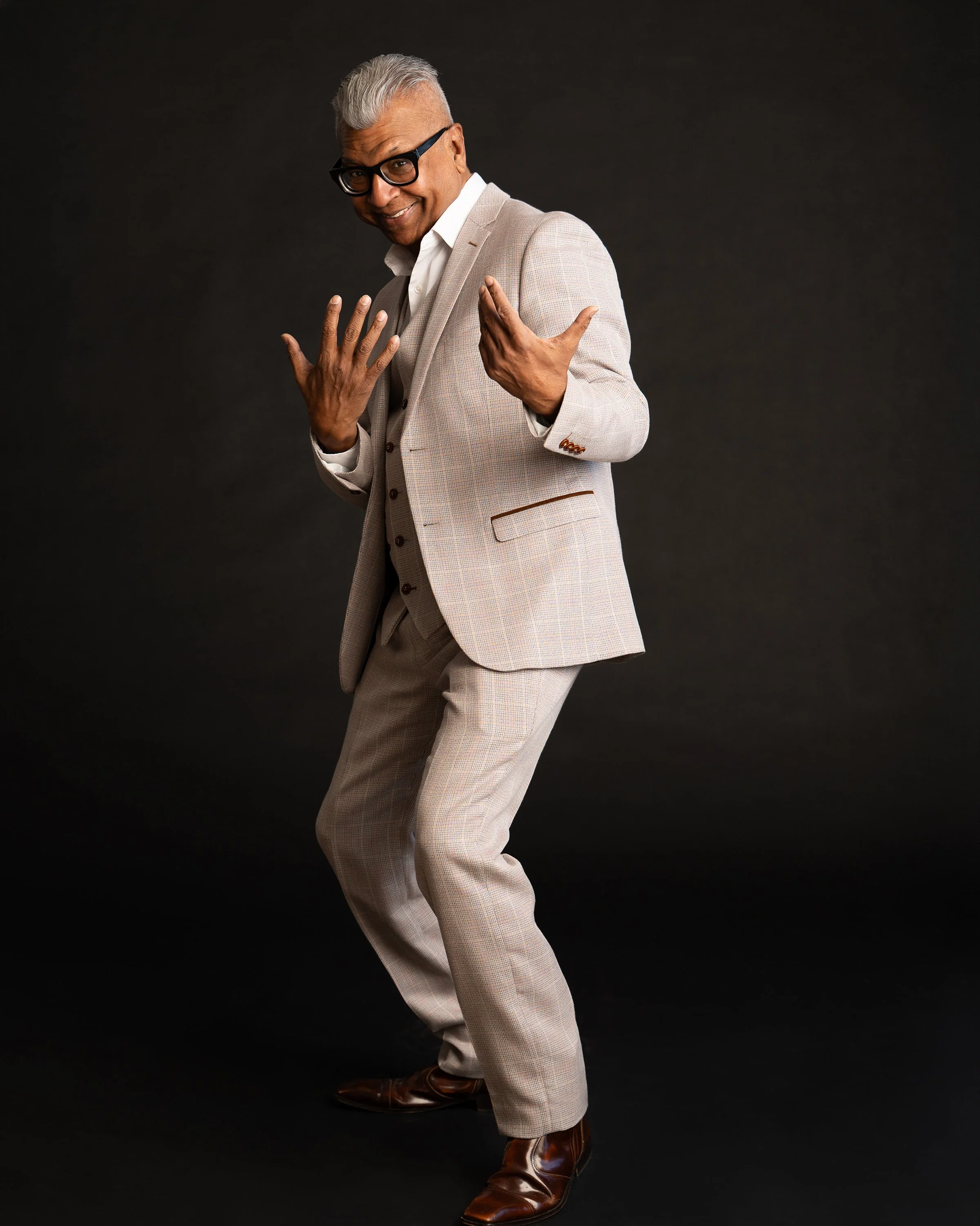 A cheerful man with gray hair, glasses, and dark skin poses confidently against a black background. He's wearing a light-colored plaid suit, white shirt, and brown boots, with one hand on his chest and the other pointing outward.