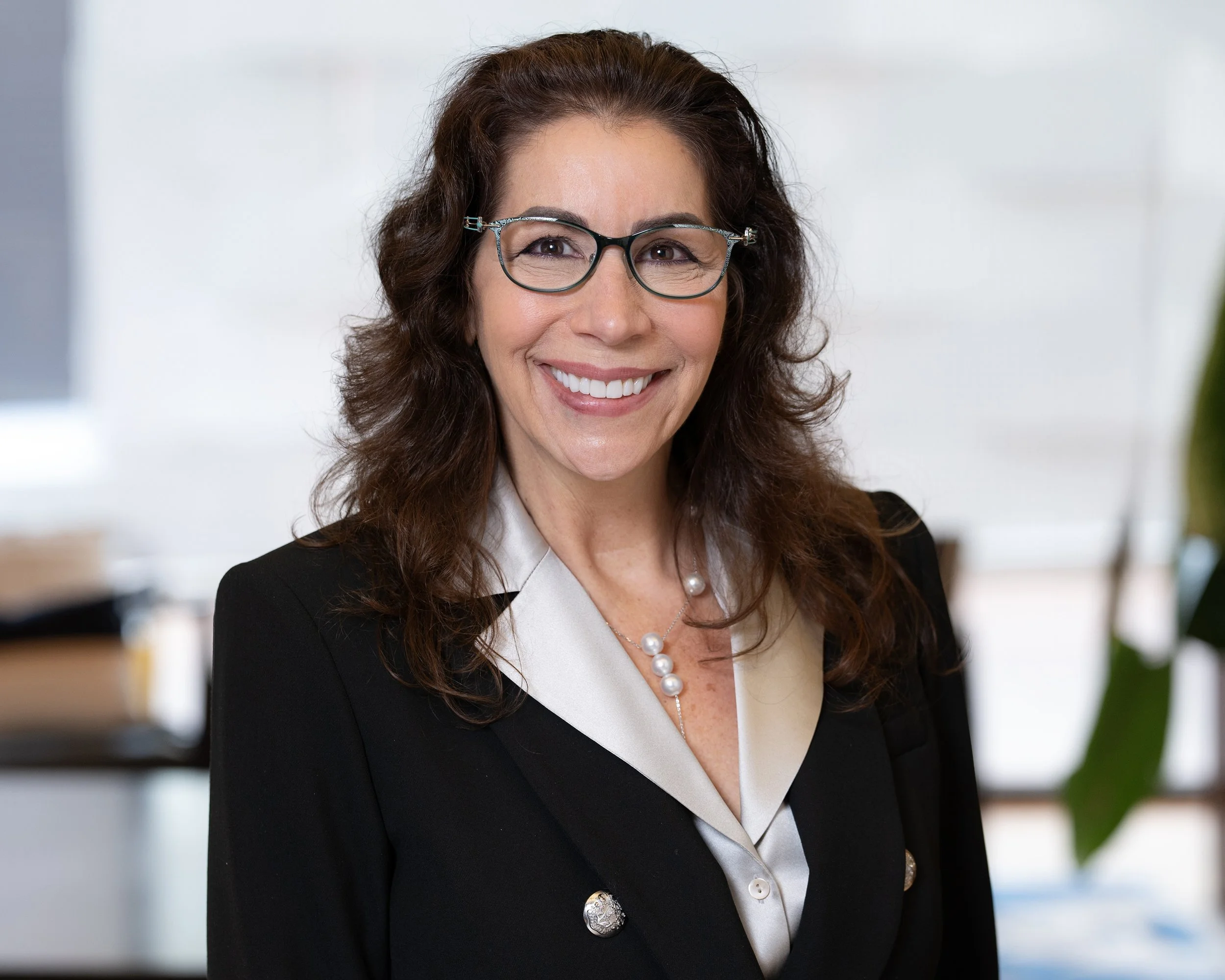 A woman with long, curly brown hair wearing glasses, a black blazer, and pearl jewelry smiling at the camera in an office setting.