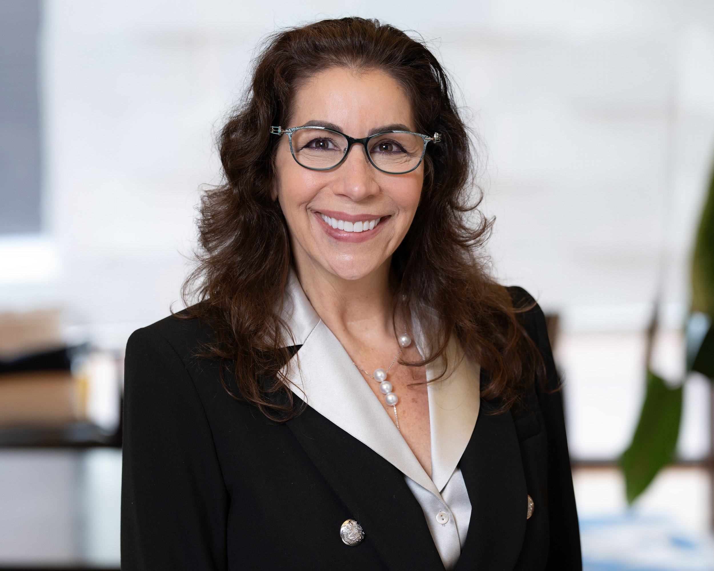 A woman with long, curly brown hair wearing glasses, a black blazer, and pearl jewelry smiling at the camera in an office setting.