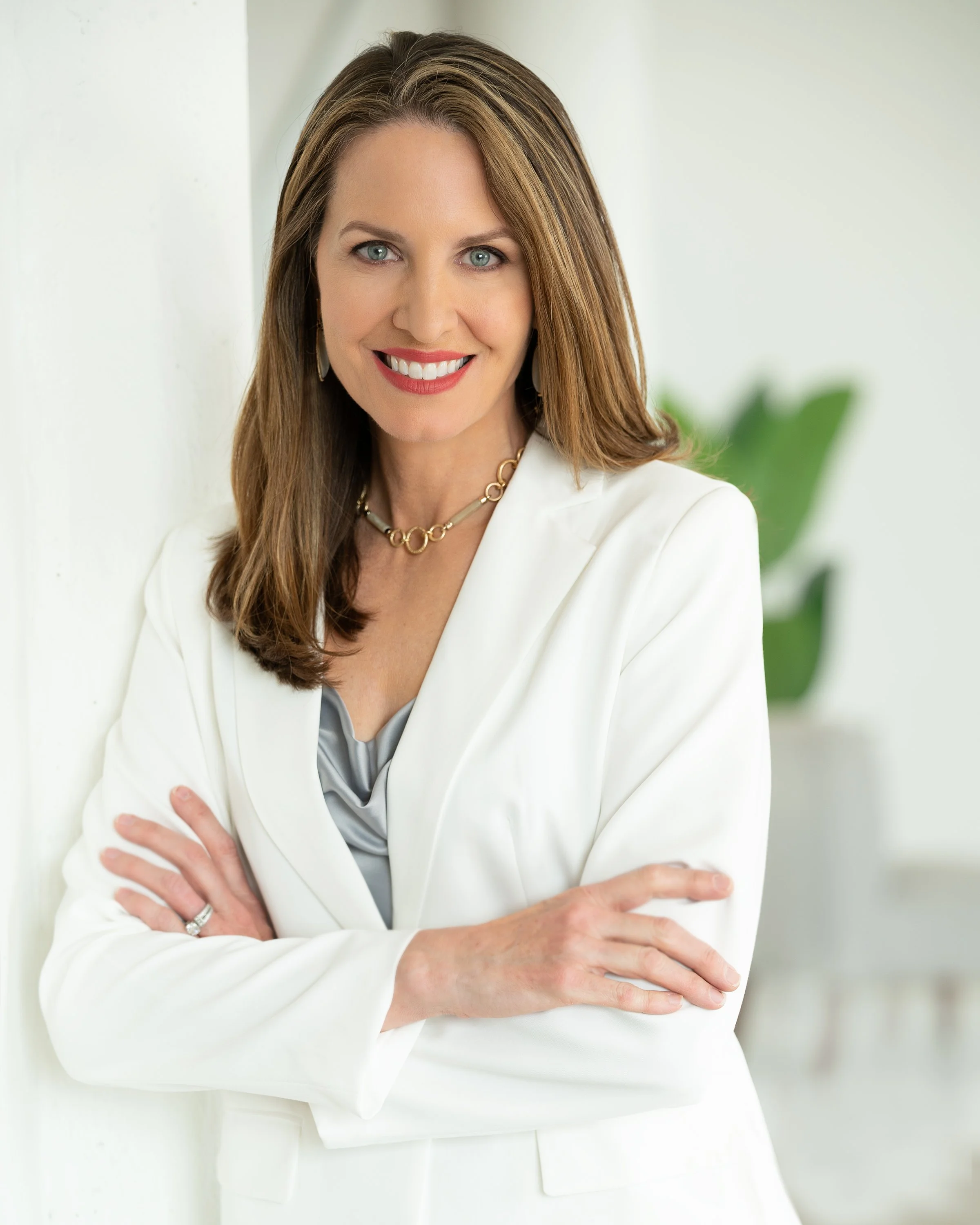 Professional woman with long brown hair, wearing a white blazer, gold necklace, and earrings, smiling with crossed arms in a bright office setting.