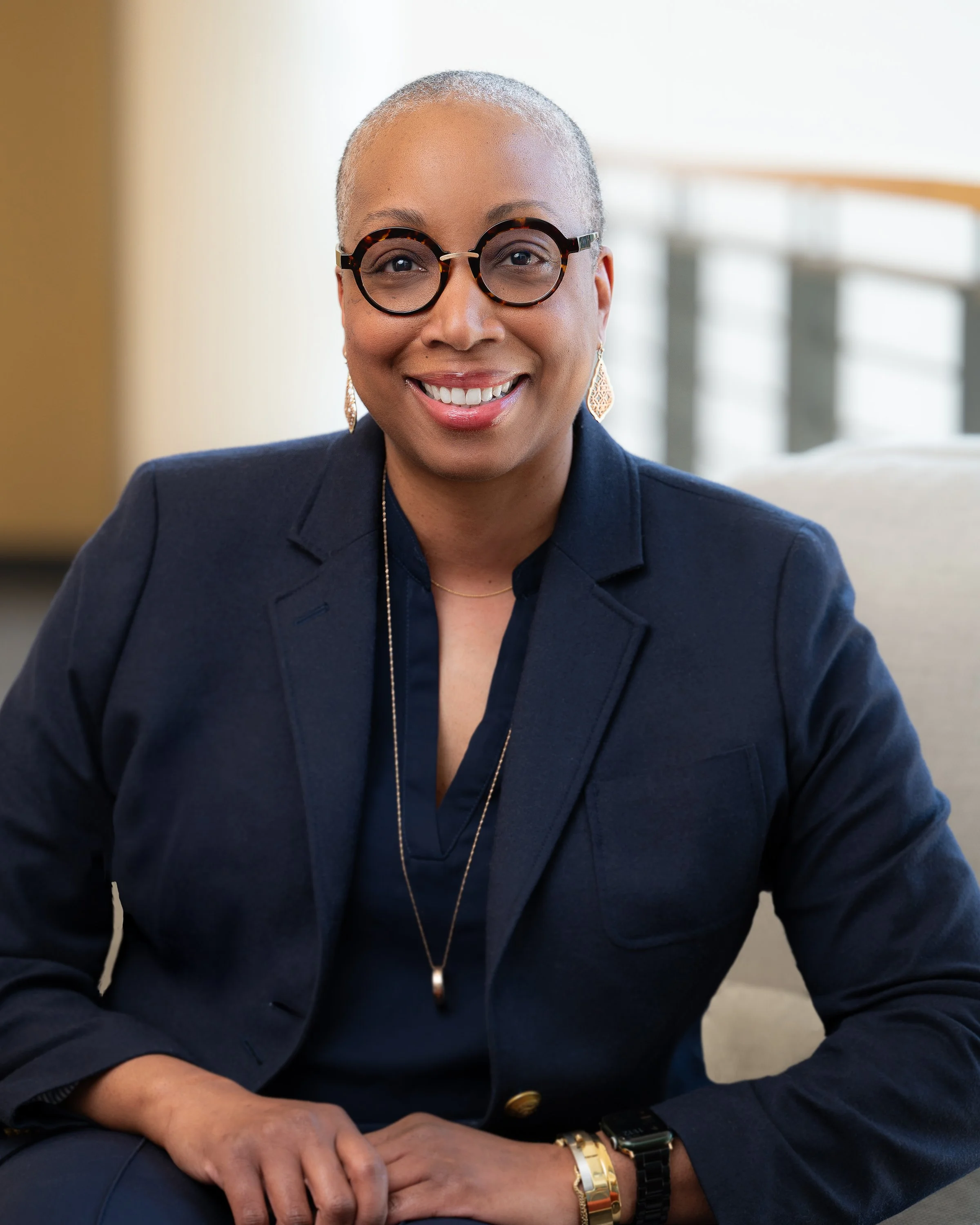 A smiling woman with short silver hair and round glasses, wearing a dark blazer and gold jewelry, sitting indoors.