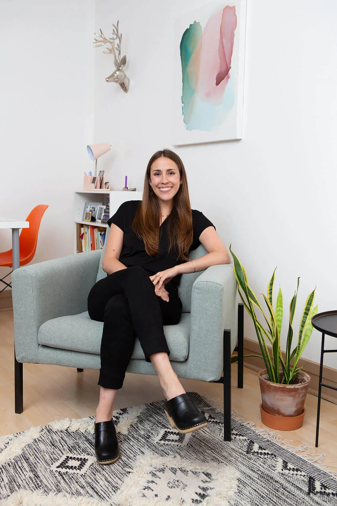 A woman with long brown hair wearing black clothing and black shoes sitting on a light gray armchair in a modern, cozy living room with houseplants, a rug, and colorful wall art.