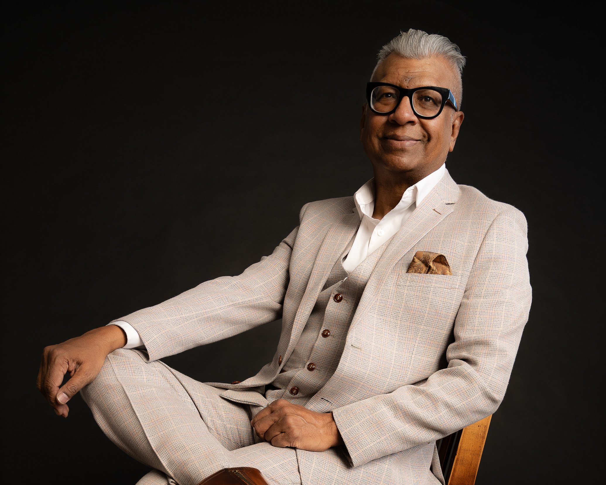A middle-aged man with gray hair and glasses sitting on a wooden chair against a black background, wearing a beige plaid suit and white shirt, smiling confidently.
