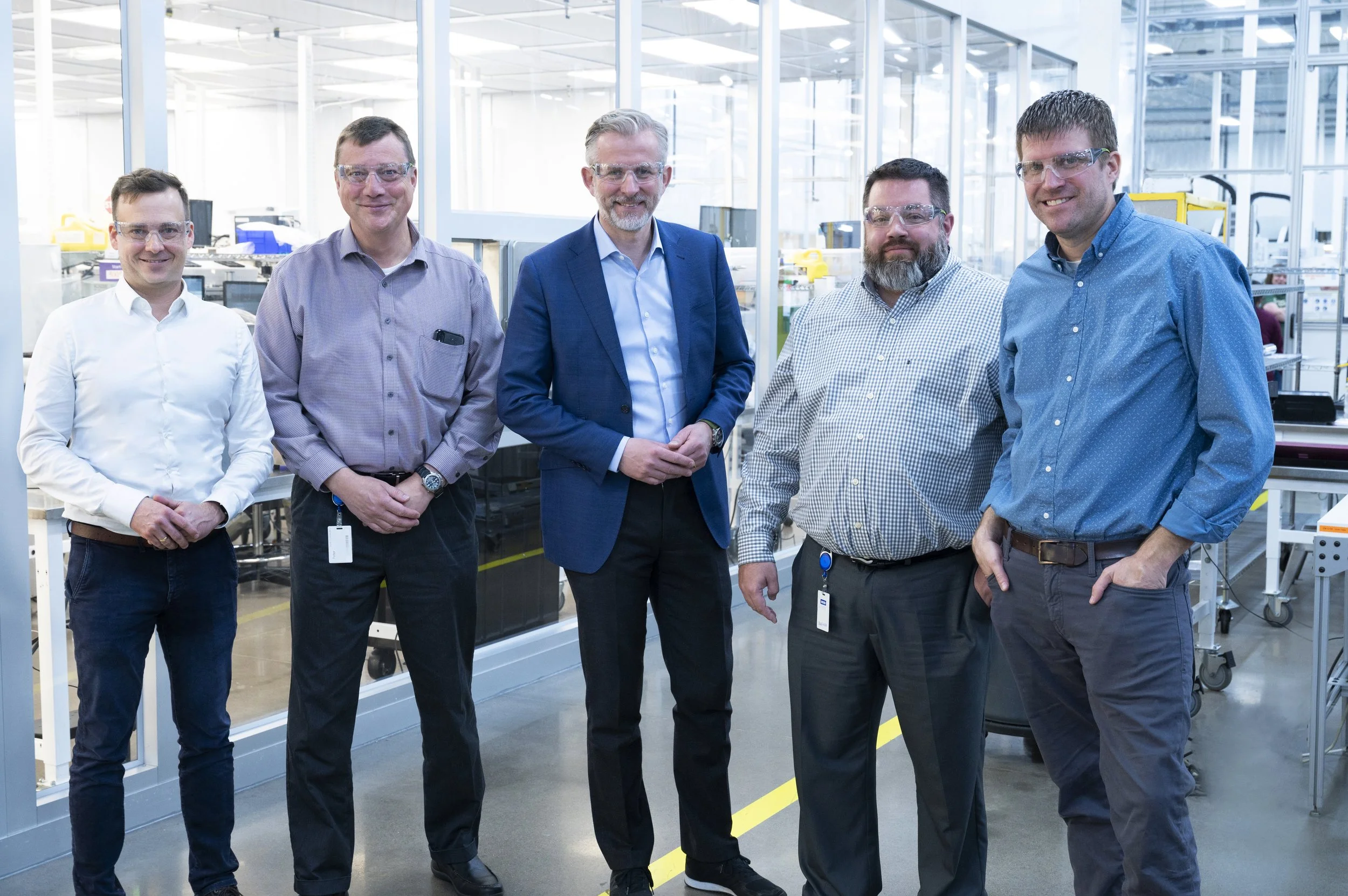 Group of five men in business casual and professional attire standing inside a laboratory or manufacturing facility with glass walls and equipment.