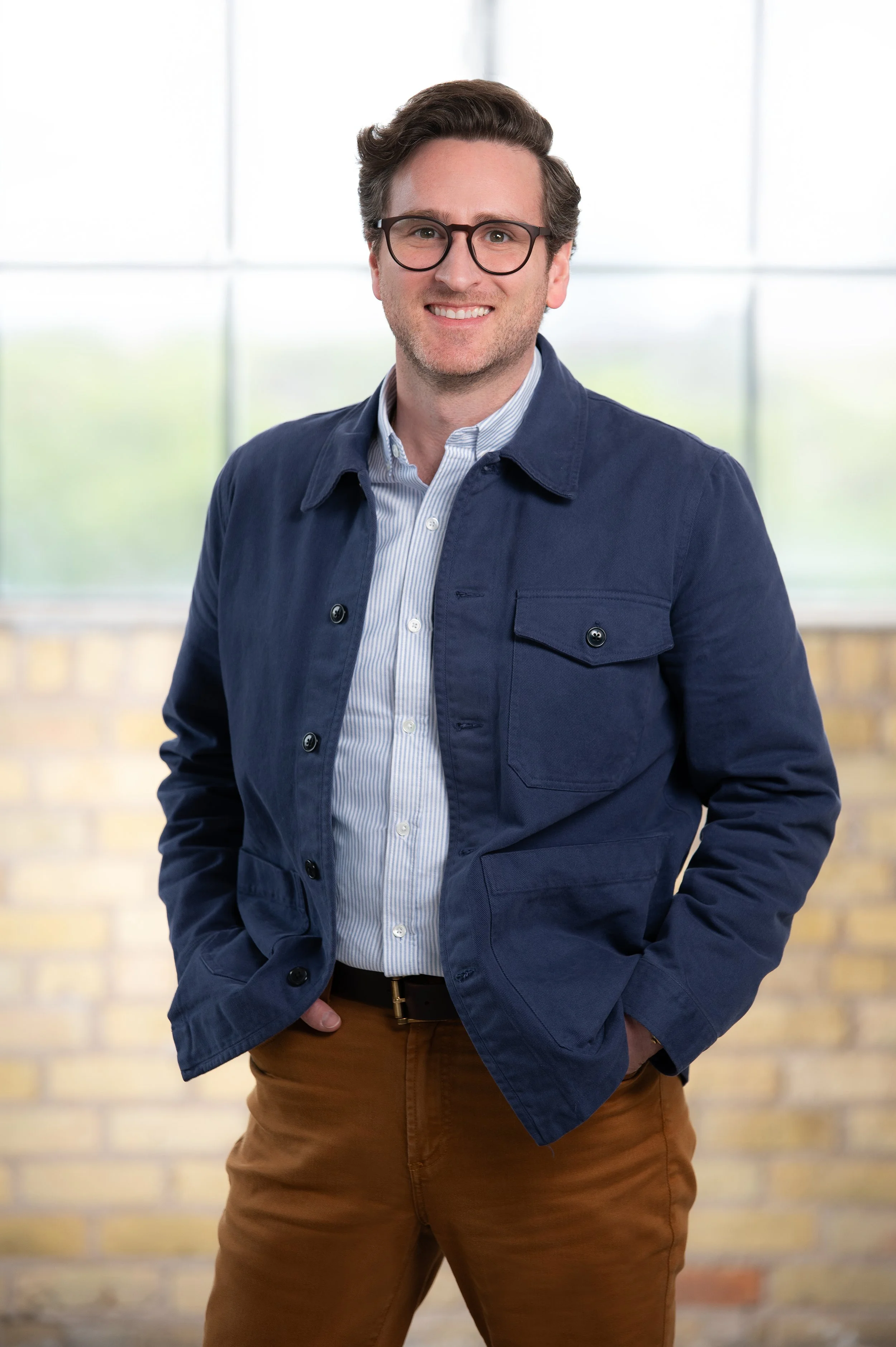 A man wearing glasses, a blue jacket, a light blue striped shirt, and brown pants, standing in front of a large window with a blurred outdoor background, smiling with his hands in his pockets.