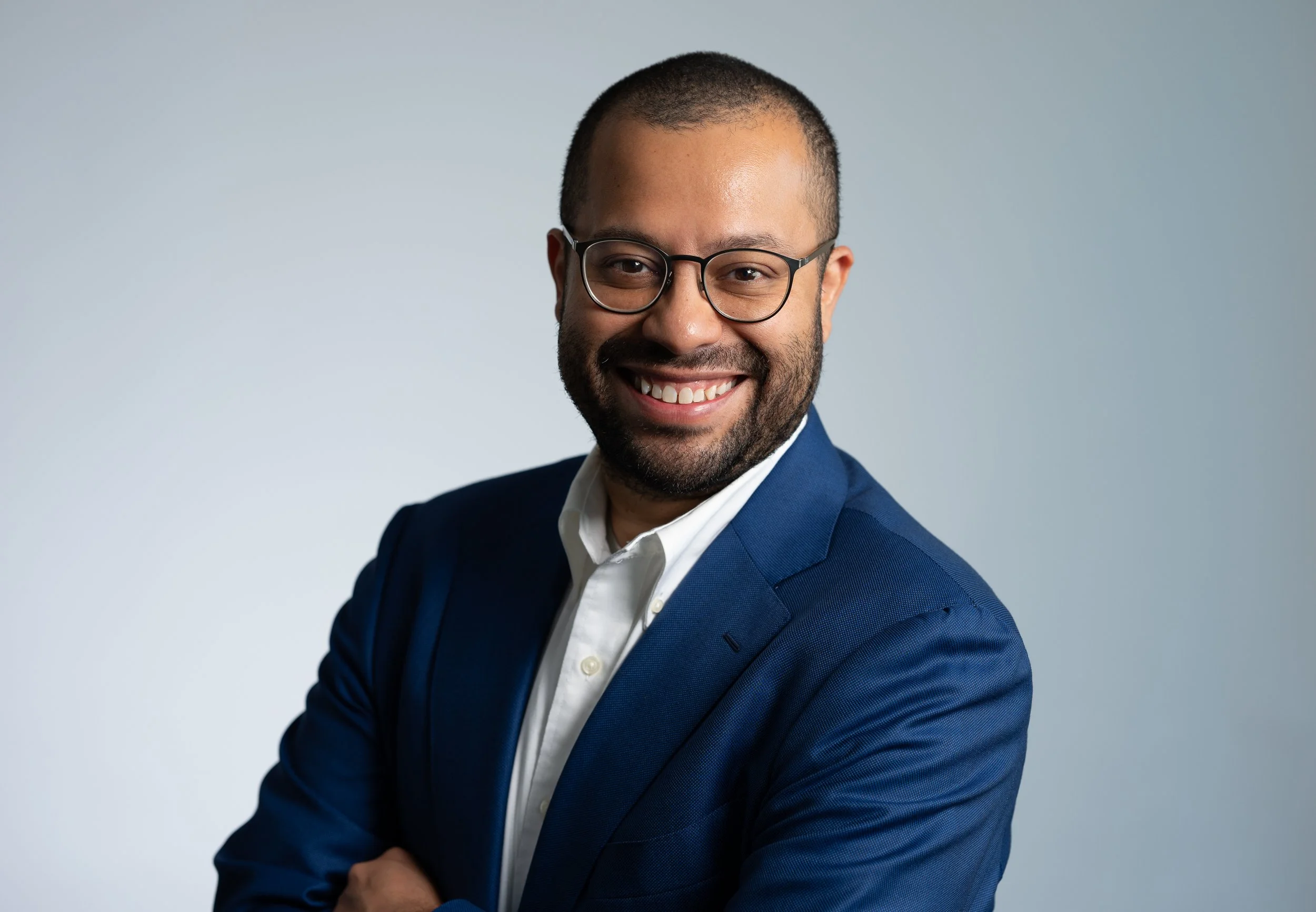 A smiling man with glasses and a beard, wearing a blue suit and white shirt, posed against a plain light gray background.
