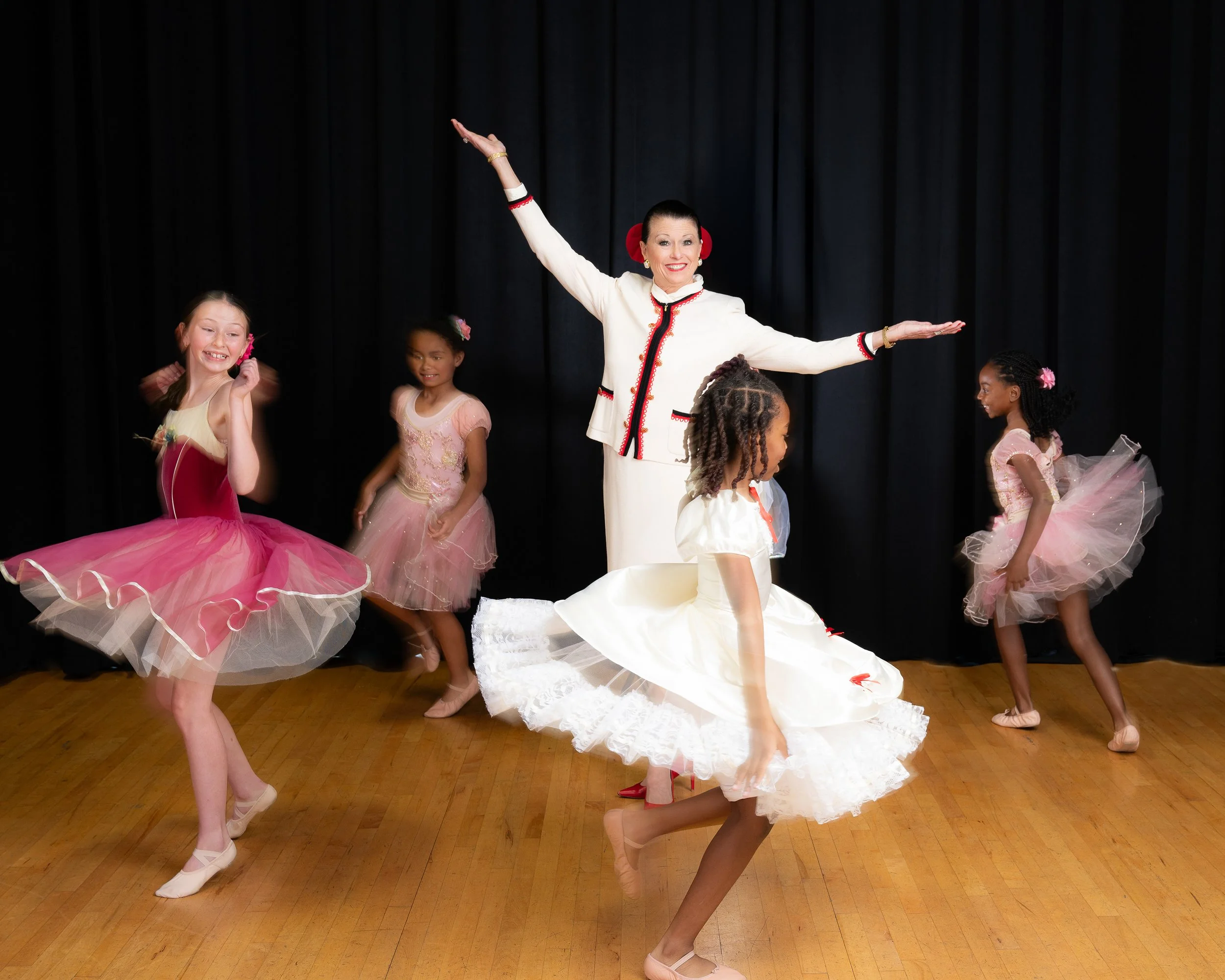 Ballet dancers performing on stage, the dancers are wearing pink and white tutus and ballet shoes, with a black curtain backdrop.