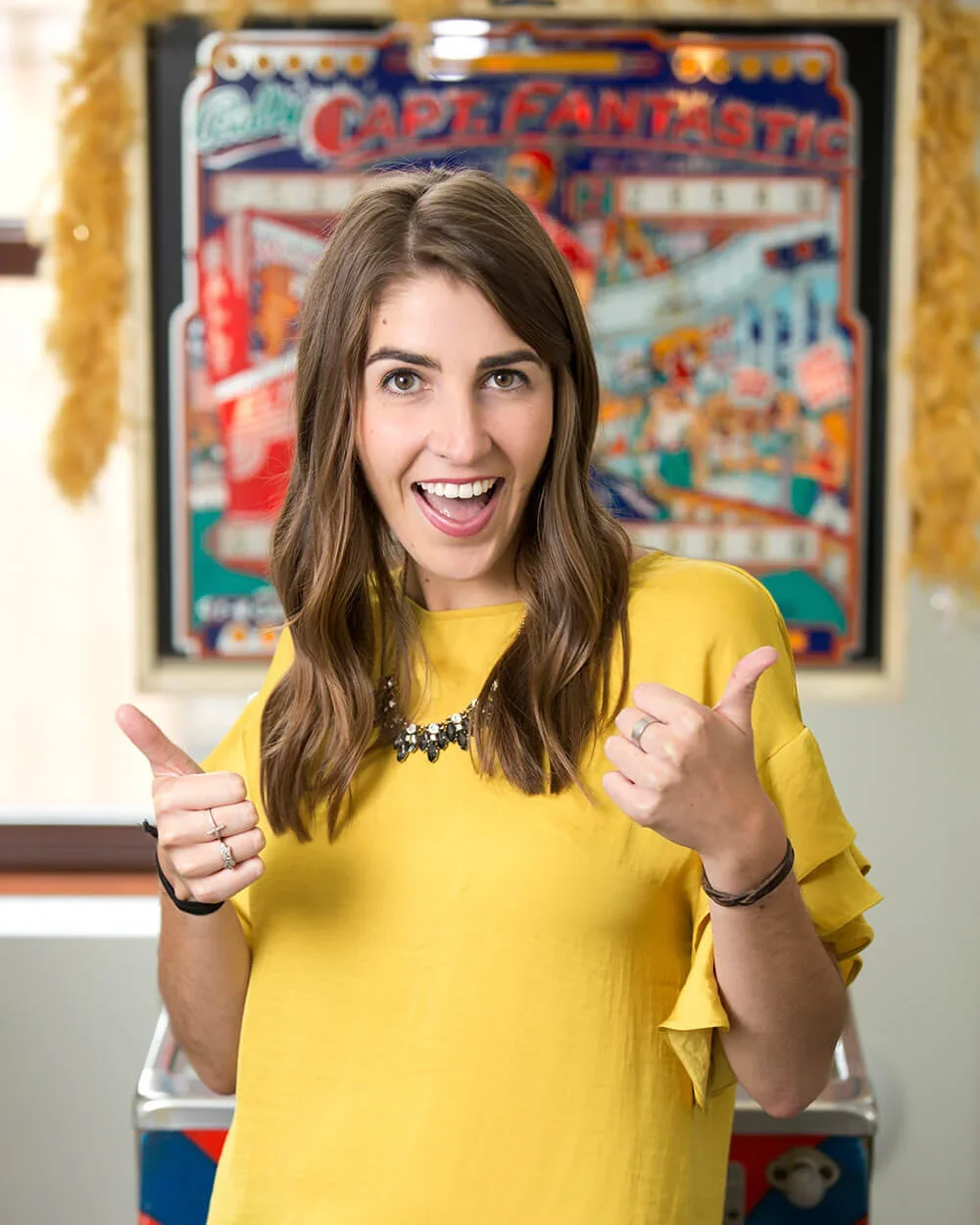 A young woman with brown hair, wearing a yellow blouse and a statement necklace, pointing and smiling at the camera in front of an colorful arcade pinball machine.