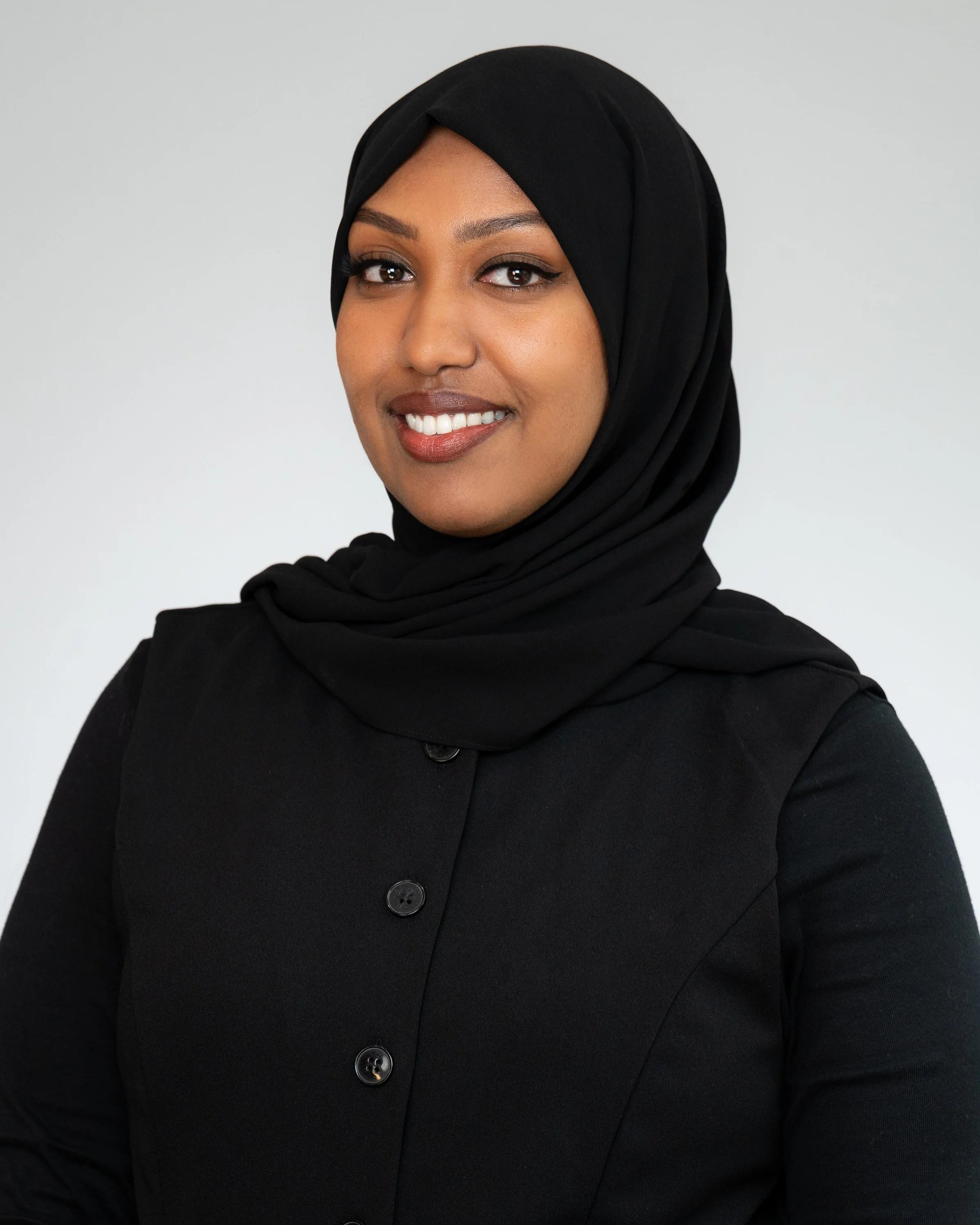 A woman wearing a black hijab and black button-up shirt, smiling, against a plain light gray background.