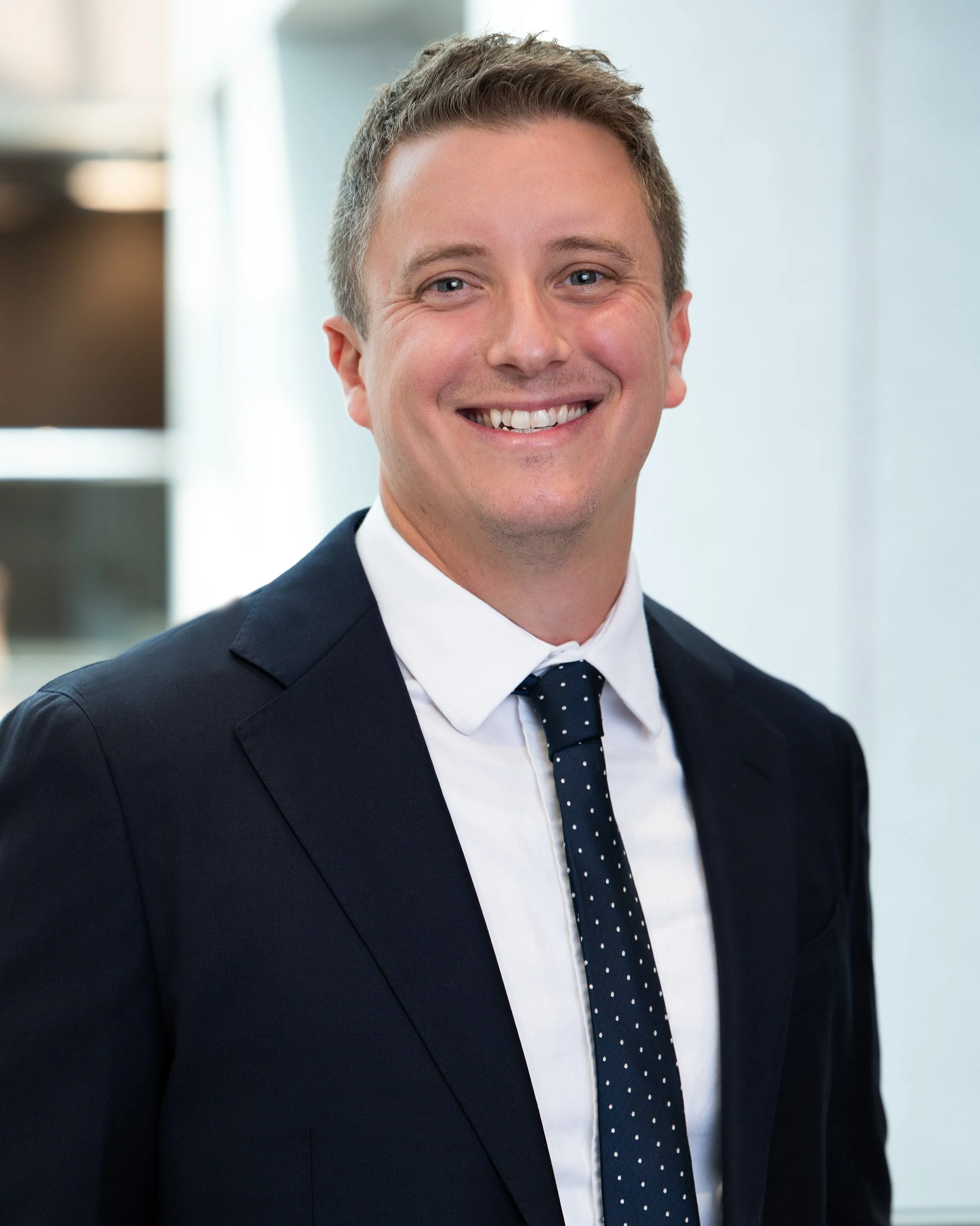 A smiling man in a dark suit, white shirt, and navy polka dot tie, standing in an office environment.