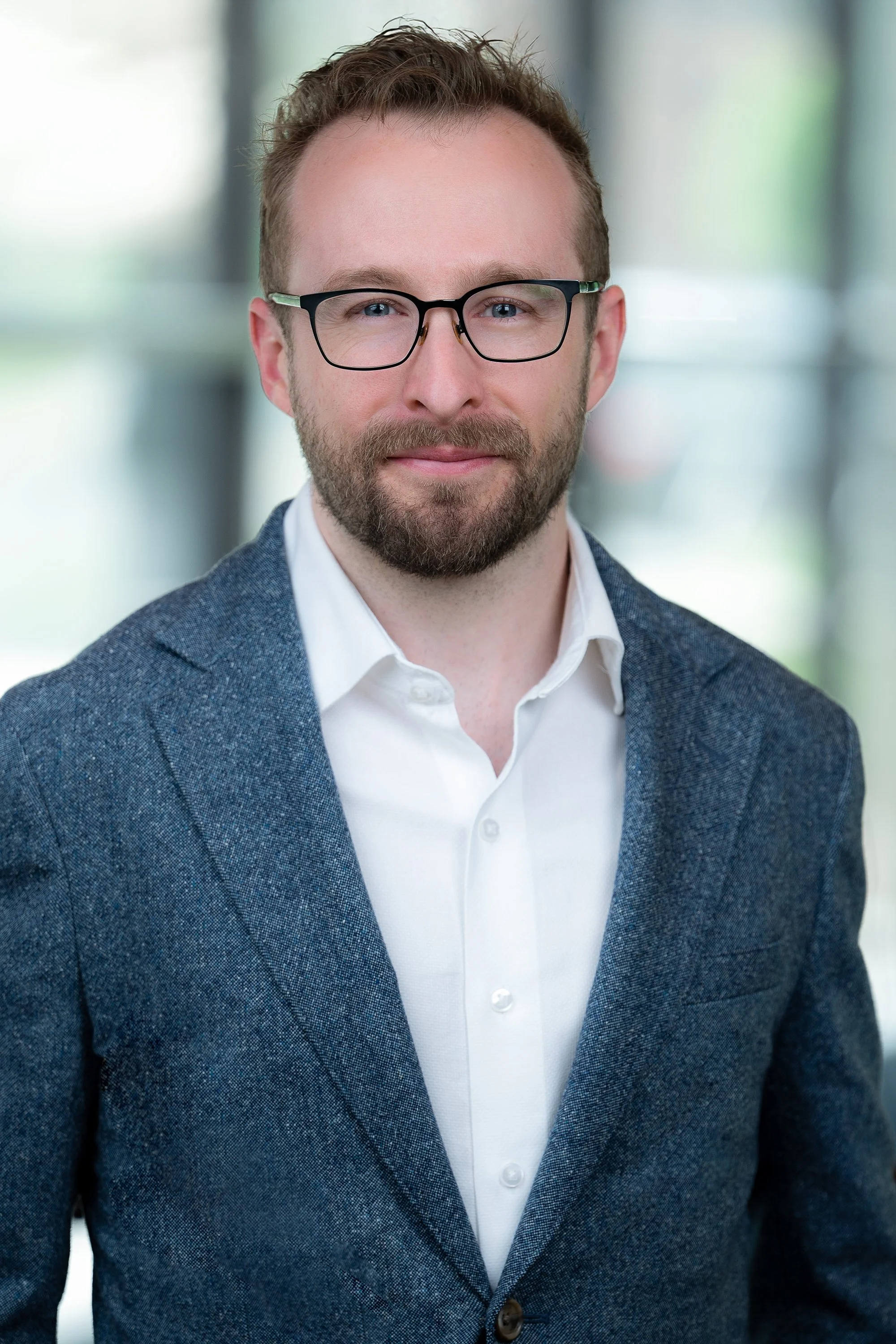 Portrait of a man with light skin, short light brown hair, trimmed beard, wearing glasses, white shirt, and blue blazer, smiling, in a modern indoor setting with blurred background.