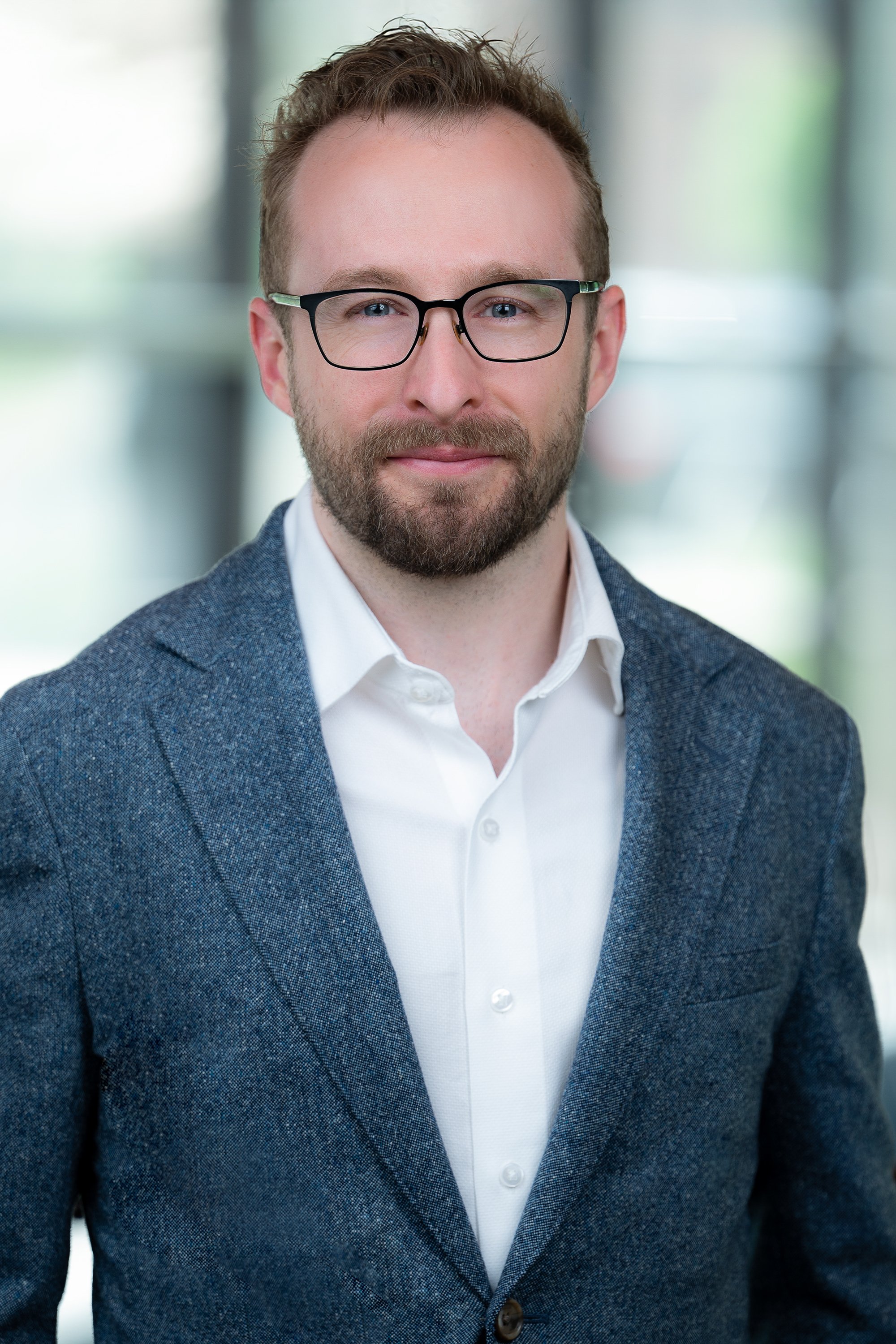 Portrait of a man with light skin, short light brown hair, trimmed beard, wearing glasses, white shirt, and blue blazer, smiling, in a modern indoor setting with blurred background.