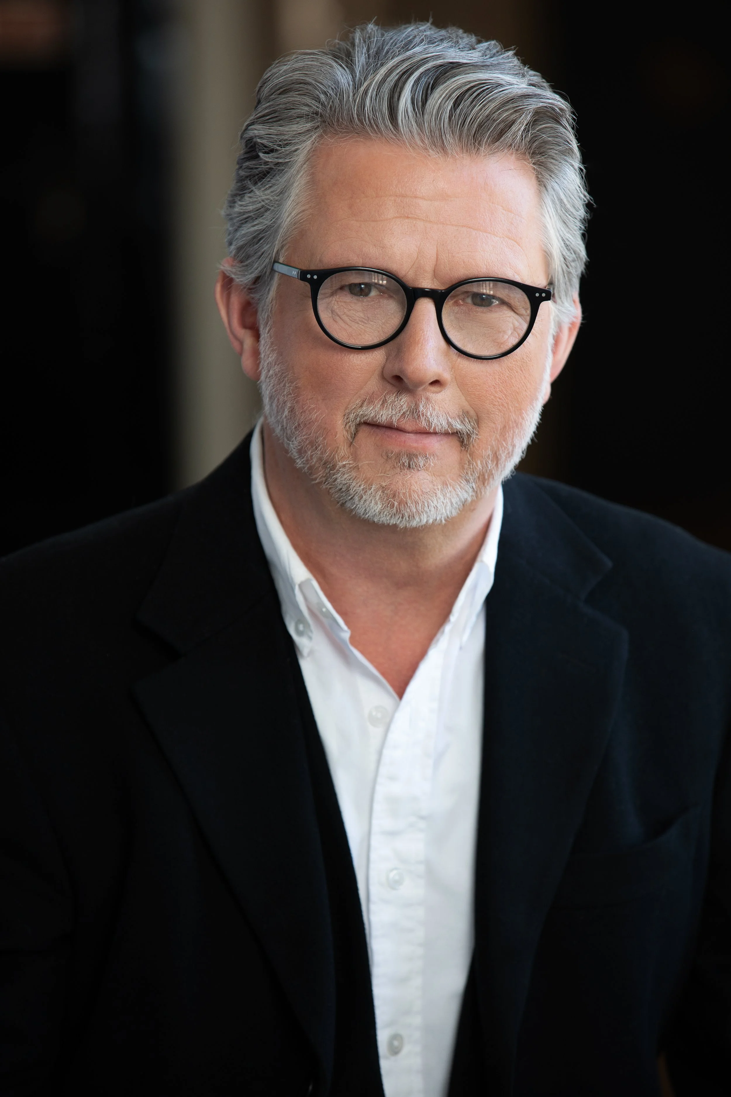A portrait of a middle-aged man with gray hair, beard, and glasses, wearing a white shirt and black blazer, against a dark background.