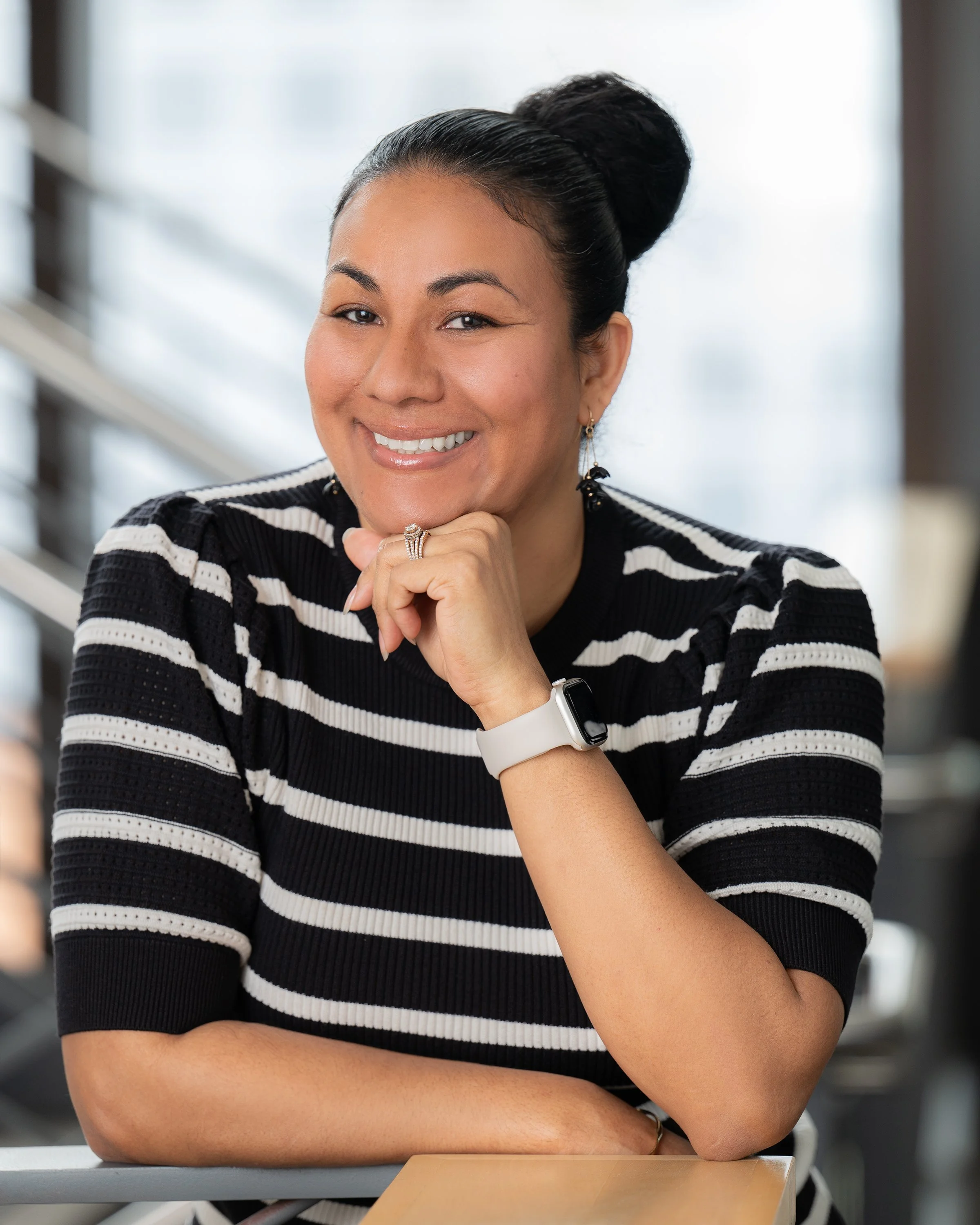 A woman with dark hair styled in a bun, wearing a black and white striped sweater, sitting at a table in an indoor setting, smiling with her right hand resting on her chin, wearing earrings, a ring, and a smartwatch.