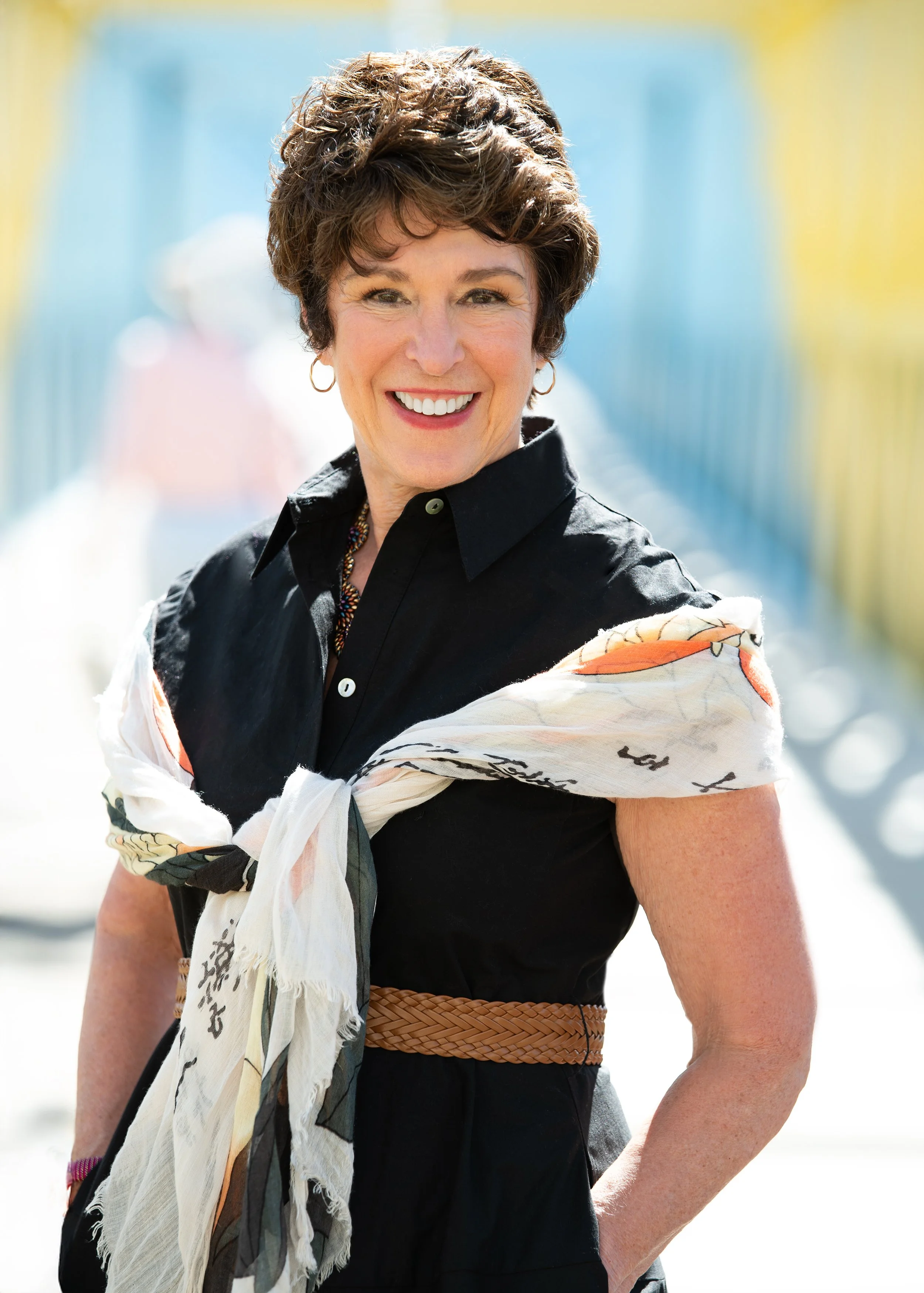 A woman with short dark hair smiling outdoors, wearing a black shirt, woven brown belt, and a light scarf with illustrations draped over her shoulders.
