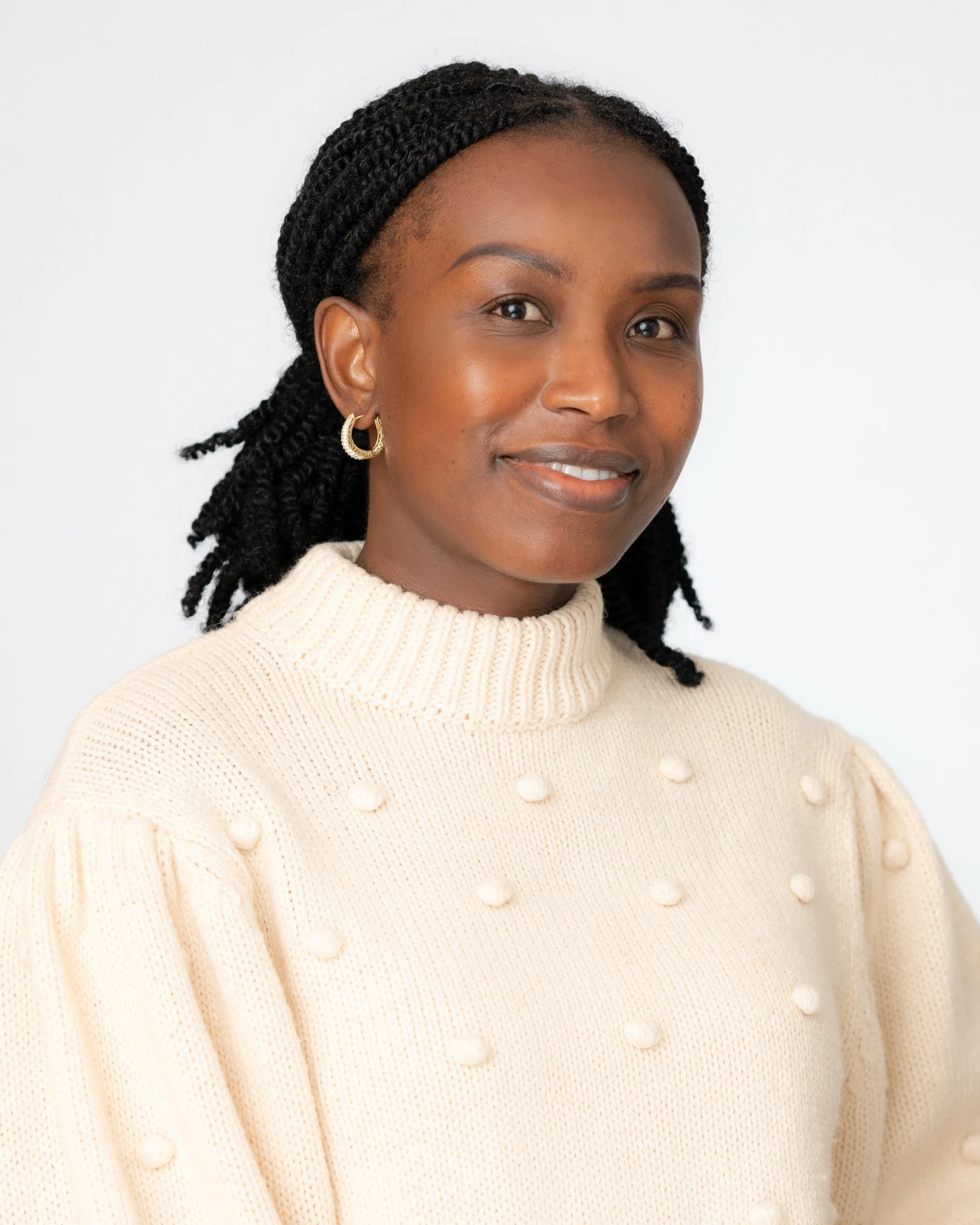 A close-up portrait of a woman with braids wearing a cream-colored sweater with pom-pom decorations and gold hoop earrings, smiling softly against a plain white background.