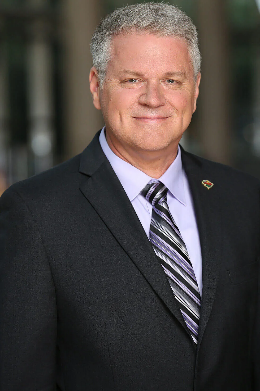 A middle-aged man with gray hair, wearing a black suit, light purple shirt, and striped purple, black, and white tie. He is smiling and standing outdoors, with an blurred background.