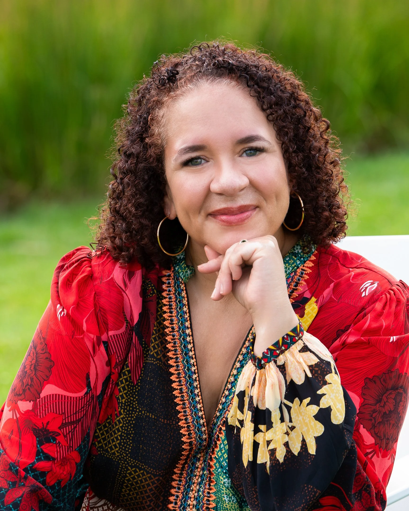 Close-up of a woman with curly brown hair, wearing gold hoop earrings and vibrant, patterned clothing in red, black, yellow, and green, outdoors with a blurred green background.