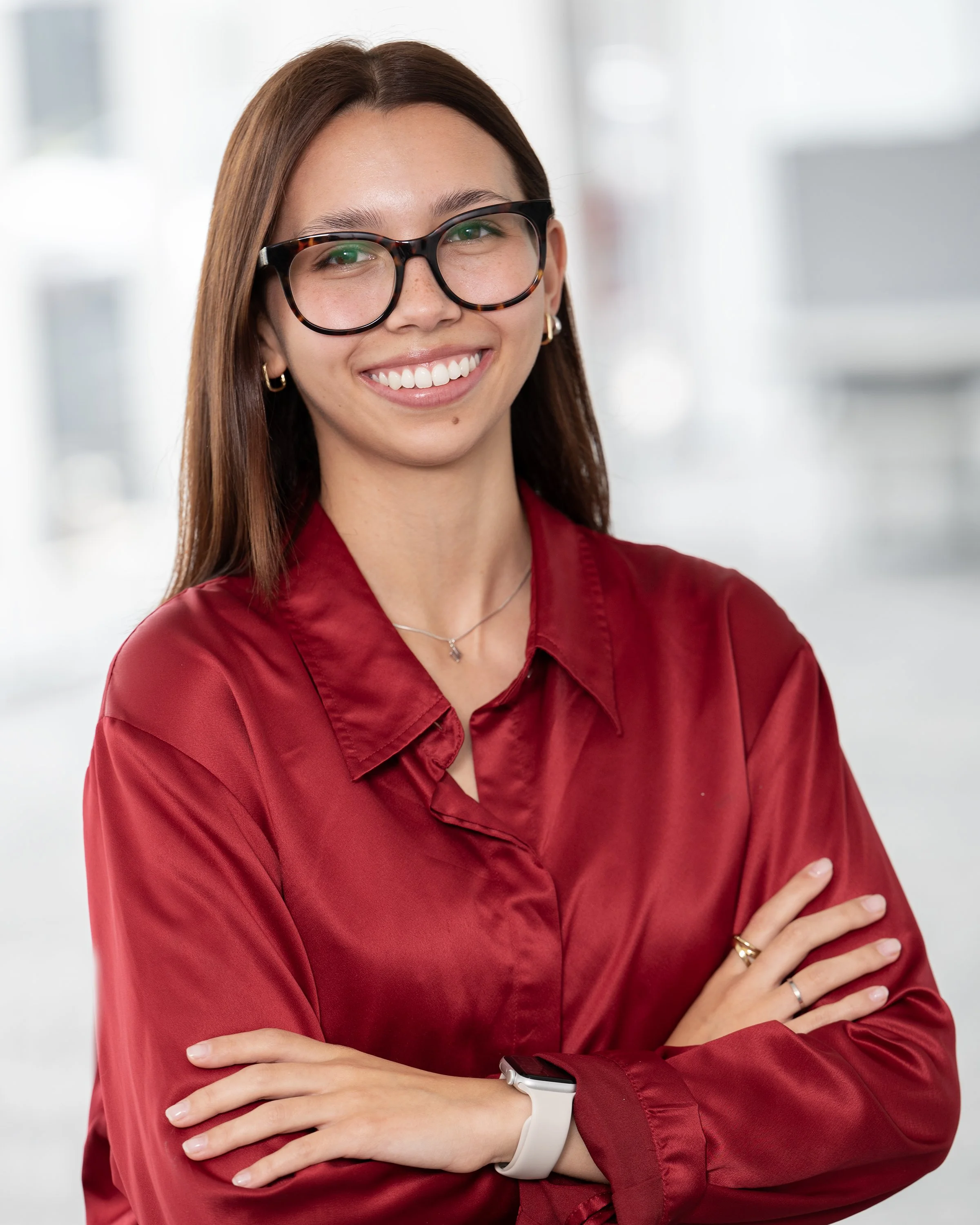 A woman with brown hair and glasses smiling, wearing a red shirt, with her arms crossed, in an office or bright indoor setting.