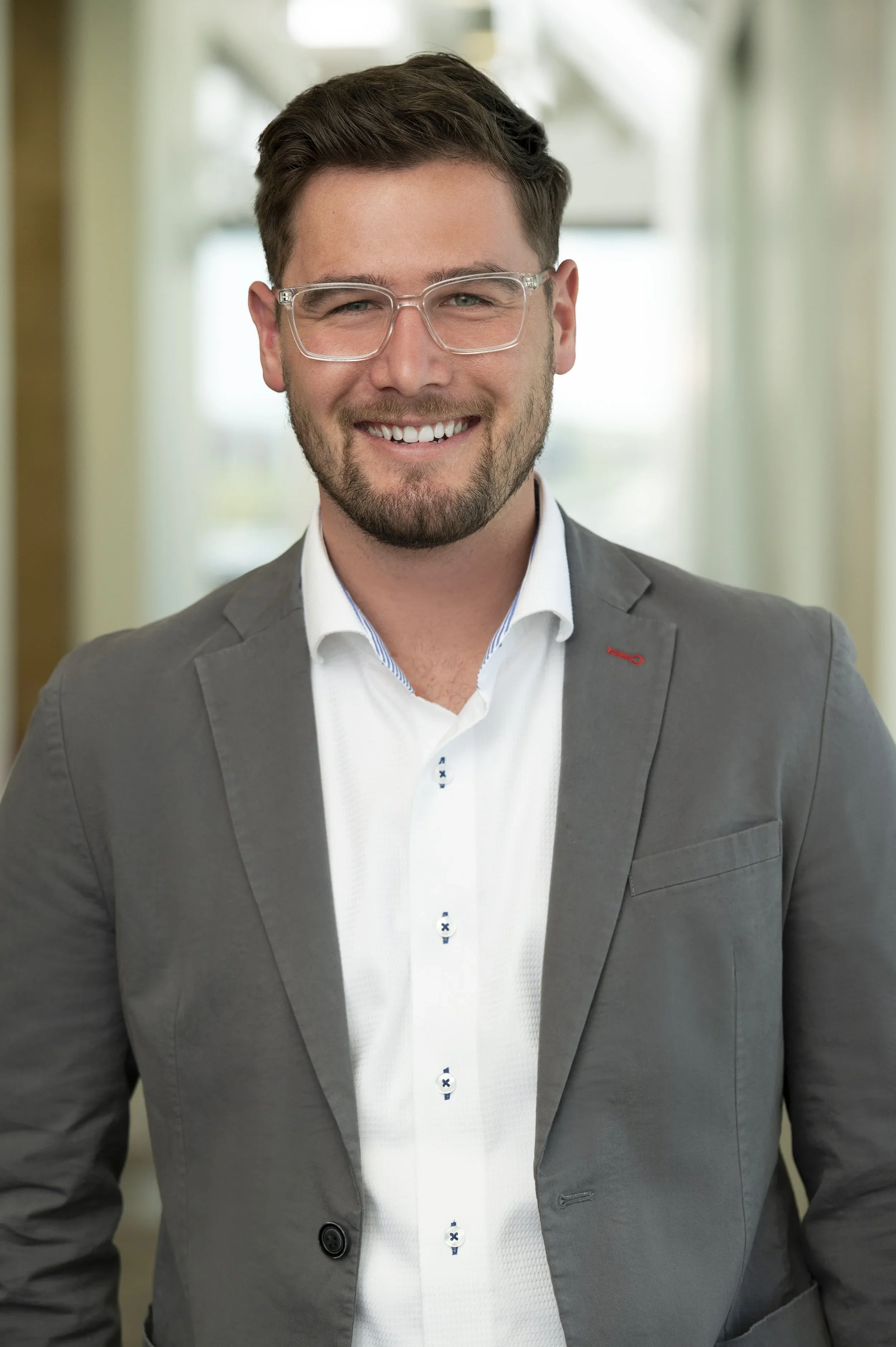 A young man with dark hair, glasses, and a beard, dressed in a gray suit and white shirt, smiling in an indoor corridor.