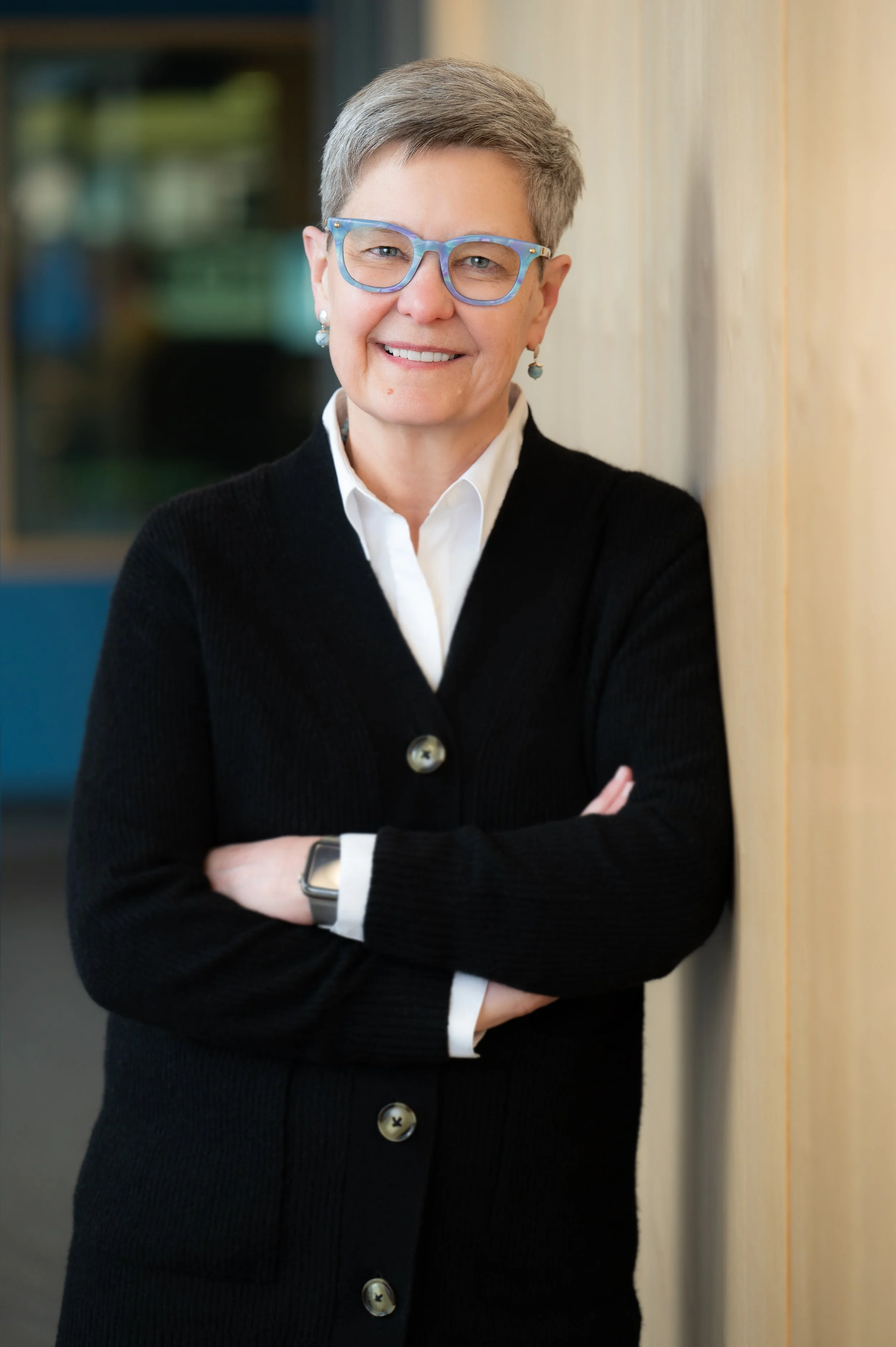 A professional woman with short gray hair, wearing blue glasses, pearl earrings, a white collared shirt, black cardigan, and watch, standing with arms crossed against a wooden wall, smiling.
