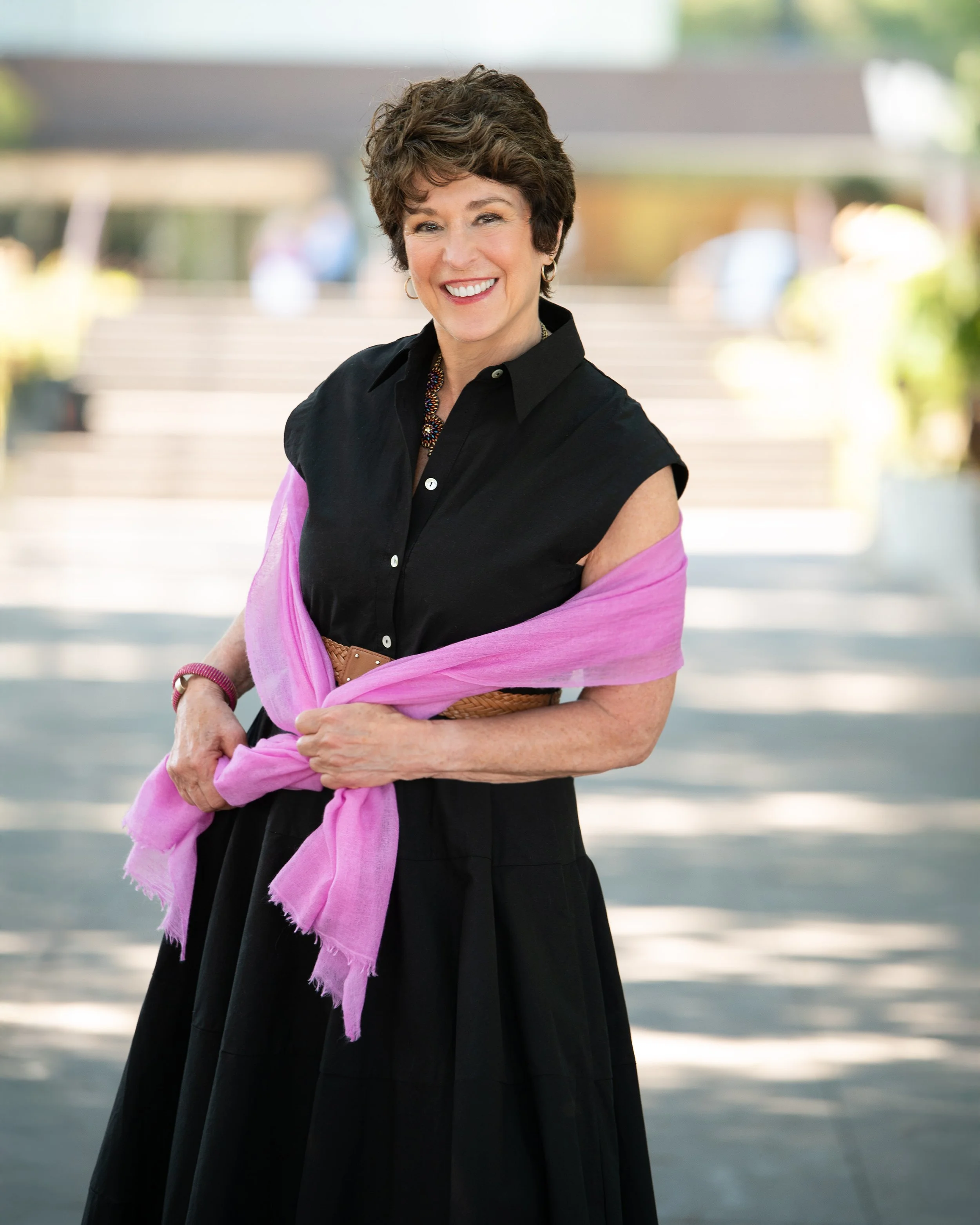 Smiling middle-aged woman with short curly hair wearing a black sleeveless shirt and a pink shawl, standing outdoors with blurred landscape background.