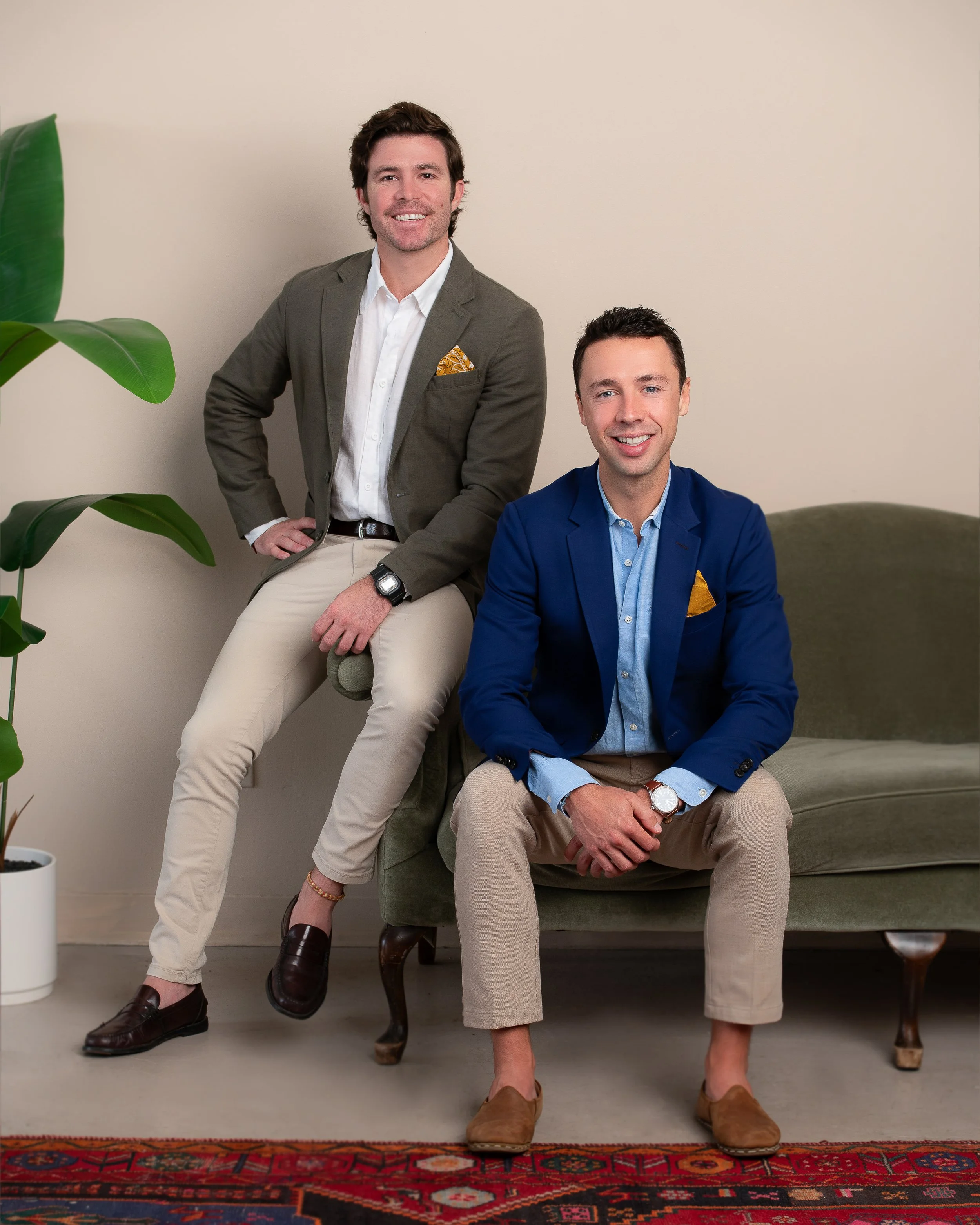 Two men in suits sitting and standing in a room with a beige wall, a potted plant on the left, a green couch on the right, and a colorful patterned rug on the floor.