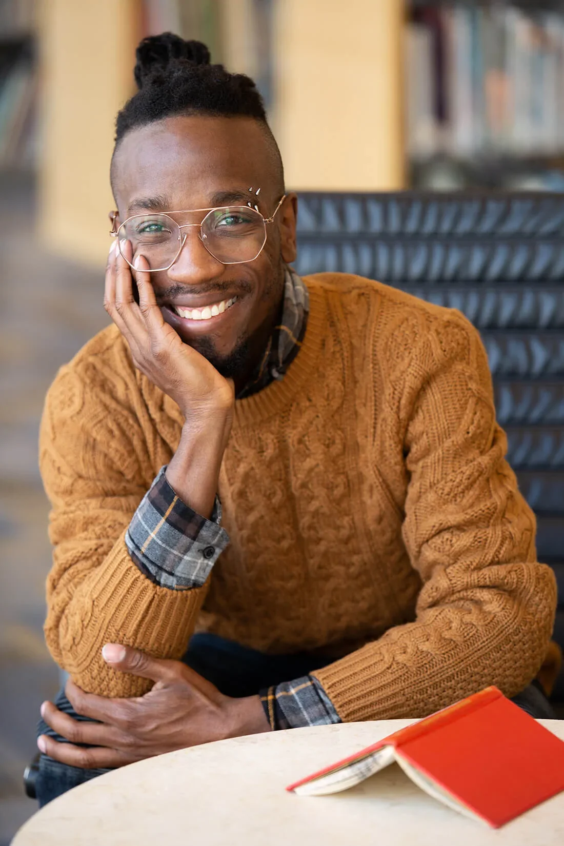 A smiling man with glasses resting his face on his hand, sitting at a table with an open red book in front of him.
