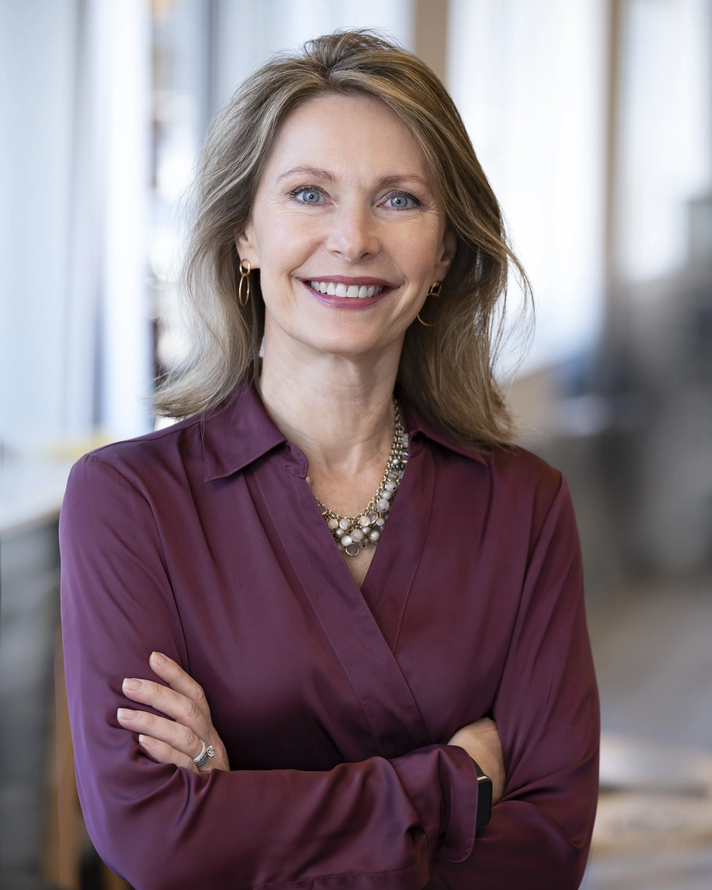 Portrait of a smiling middle-aged woman with blue eyes and light brown hair, wearing a maroon blouse, a pearl necklace, gold hoop earrings, and a ring on her finger, standing indoors near a window.