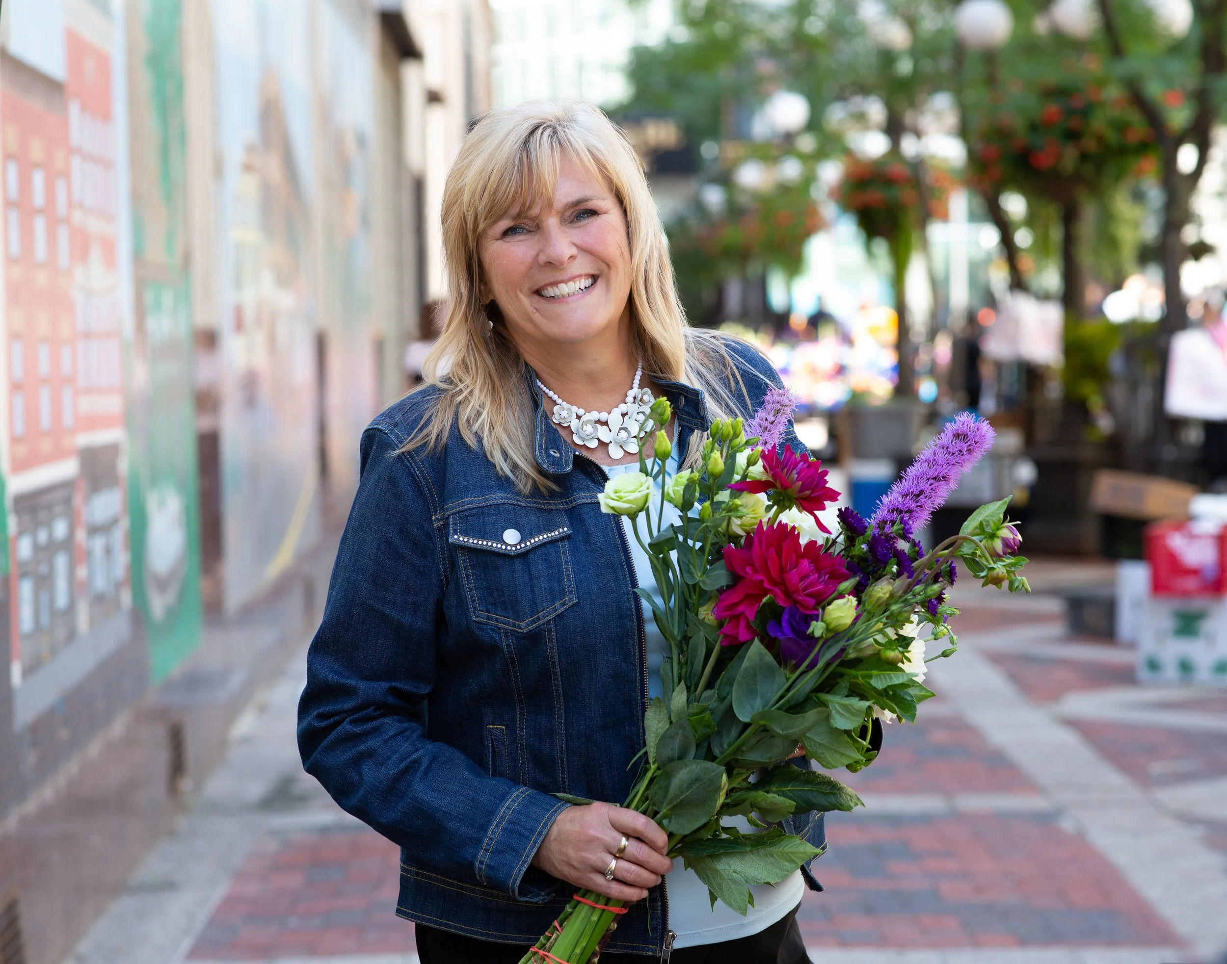 A woman with blonde hair smiling while holding a large bouquet of colorful flowers outdoors.