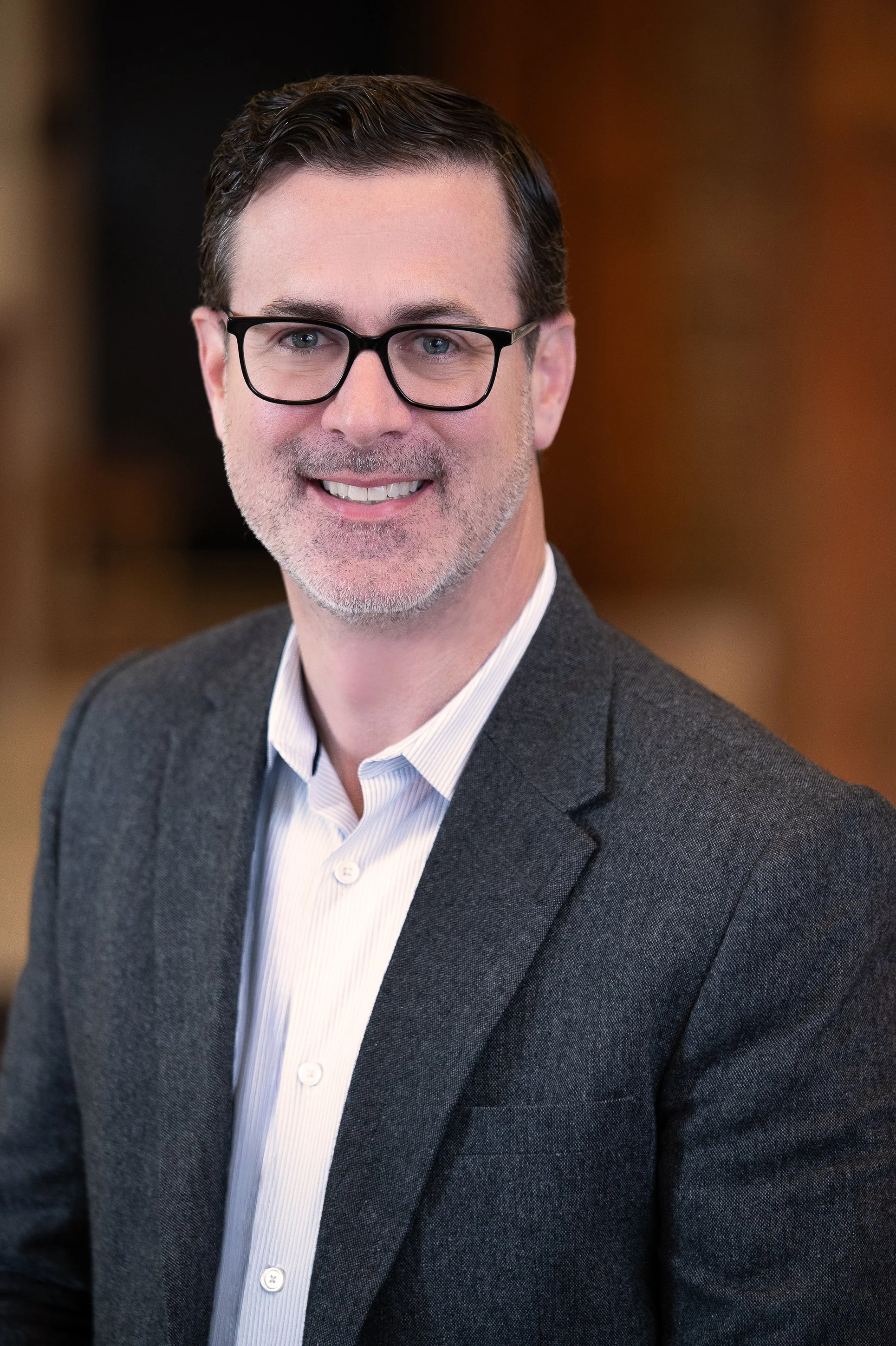 Portrait of a man wearing glasses and a gray suit, smiling in an indoor setting.