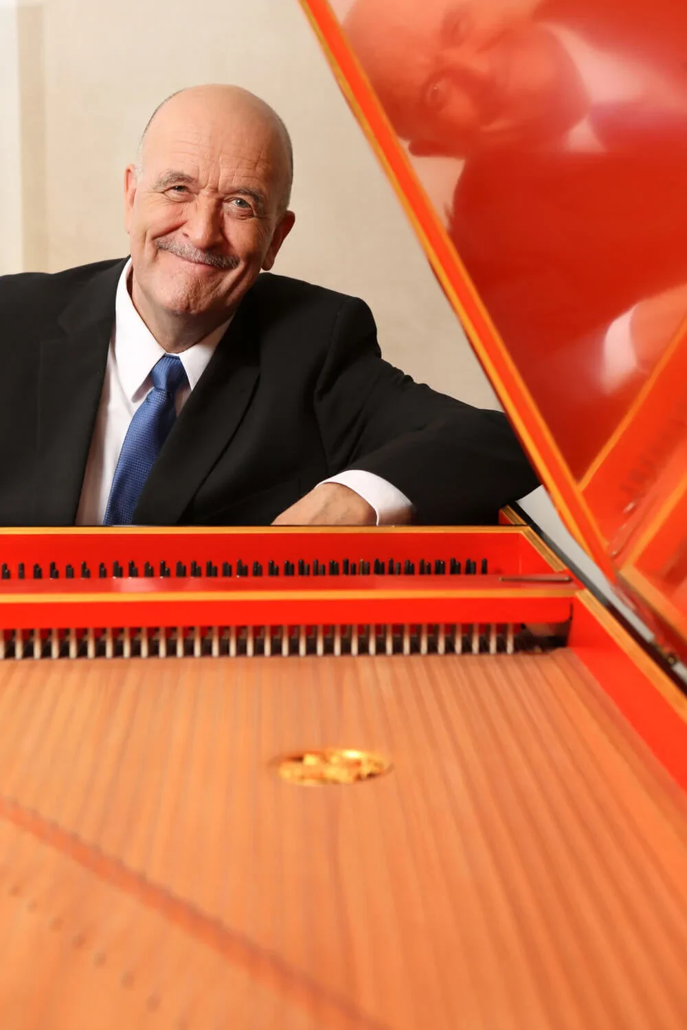 A smiling man in a black suit with white shirt and blue tie sitting at a red grand piano.