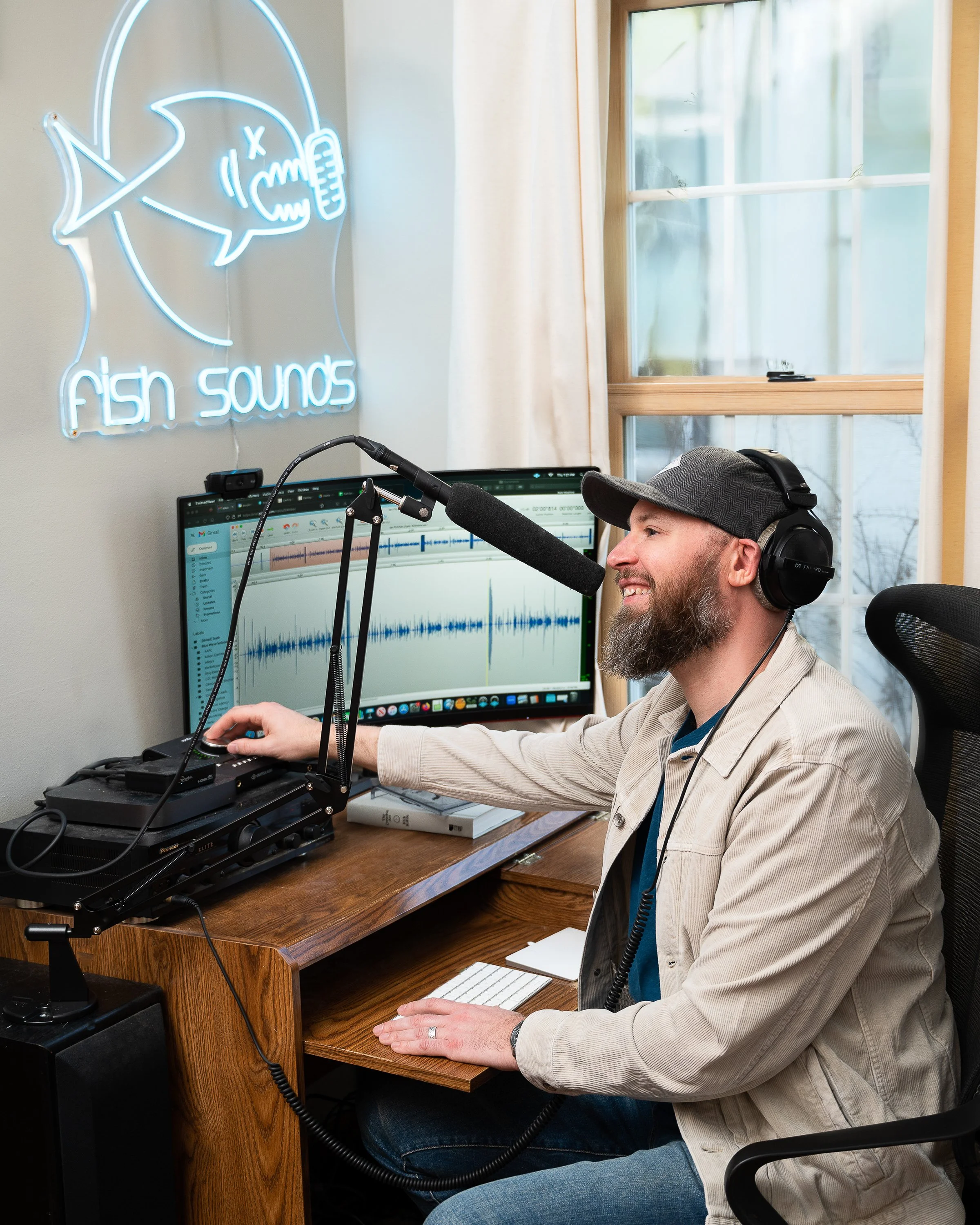 A man with a beard and wearing a gray cap and beige jacket is sitting at a wooden desk using a microphone connected to a computer, with a music editing software open on the screen. There is a neon sign on the wall that says 'fish sounds' with a fish 