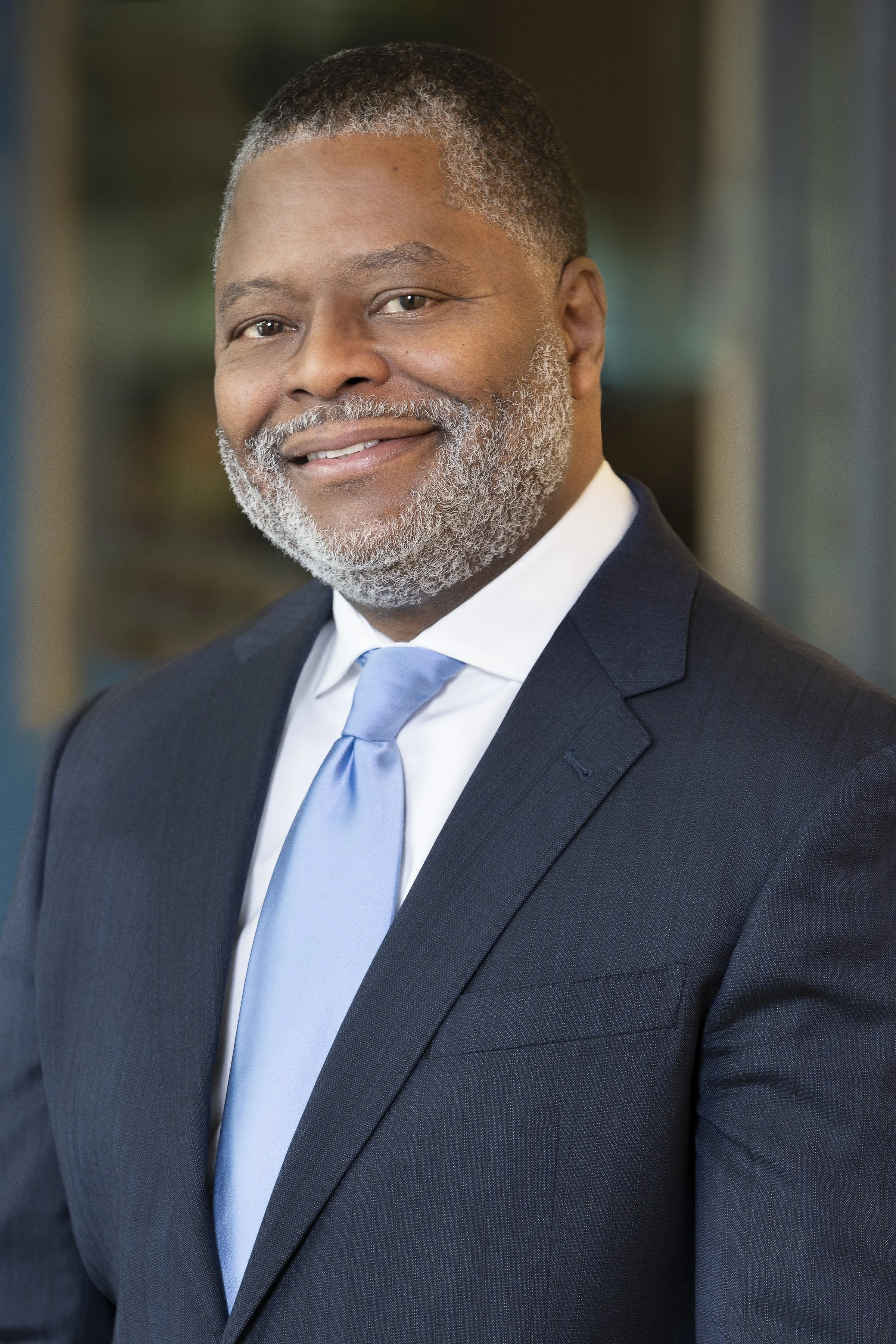 Close-up of a smiling African American man wearing a dark suit, light blue tie, and white shirt, in a professional setting.