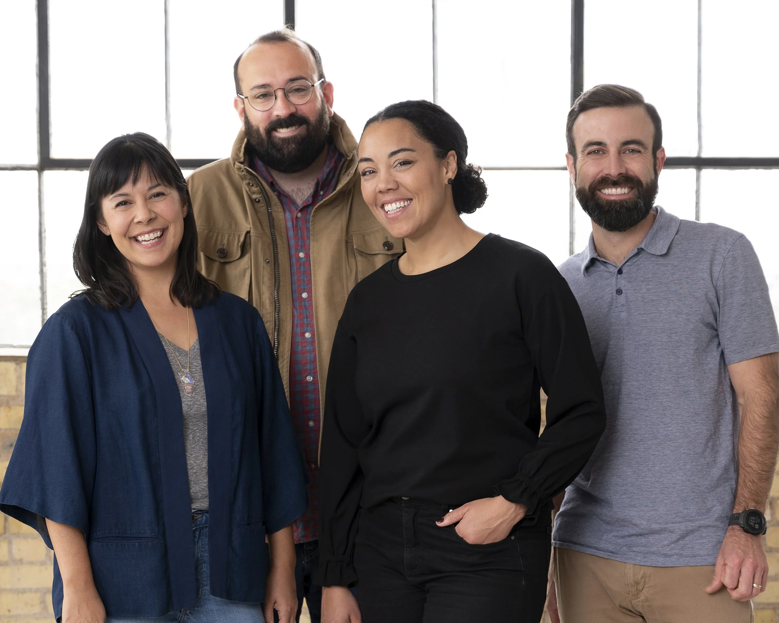 A diverse group of four smiling people standing together indoors near large windows.