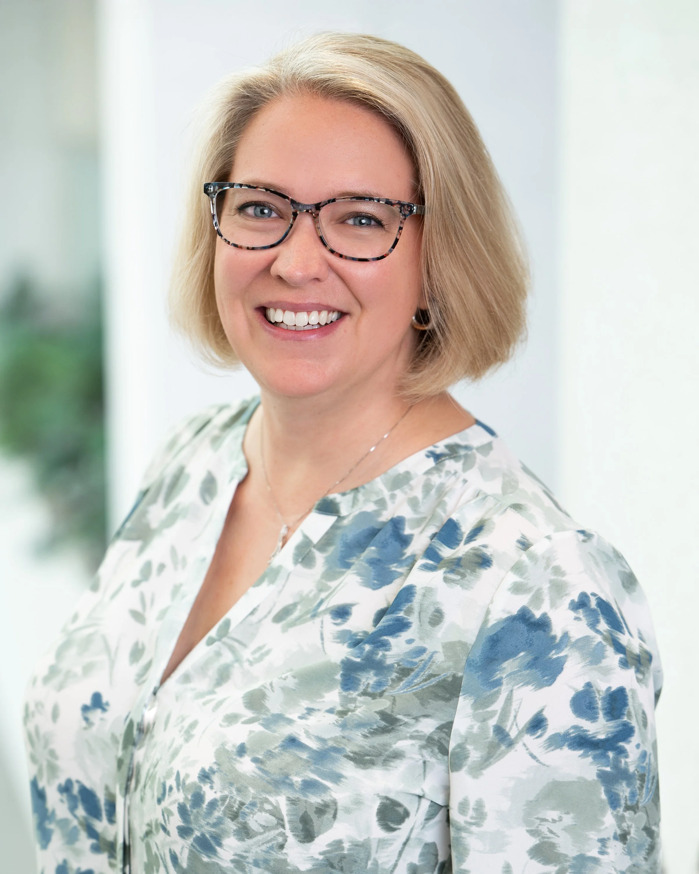 Close-up of a smiling woman with blonde hair, wearing tortoiseshell glasses and a white and blue floral blouse, standing indoors with a light-colored background.