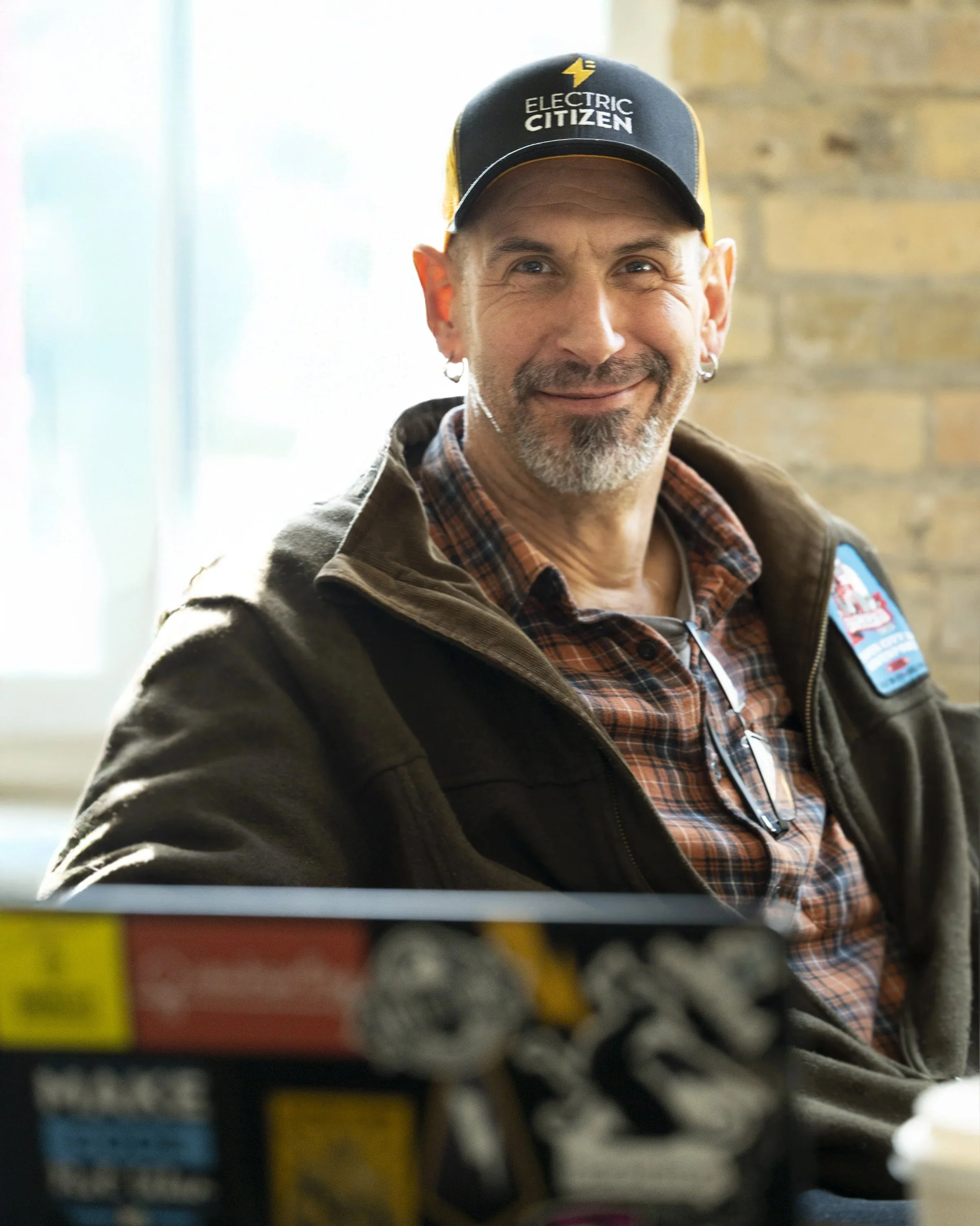 Smiling man wearing a black baseball cap with 'Electric Citizen' logo, a plaid shirt, and a brown jacket, sitting in a store with shelves full of colorful products.