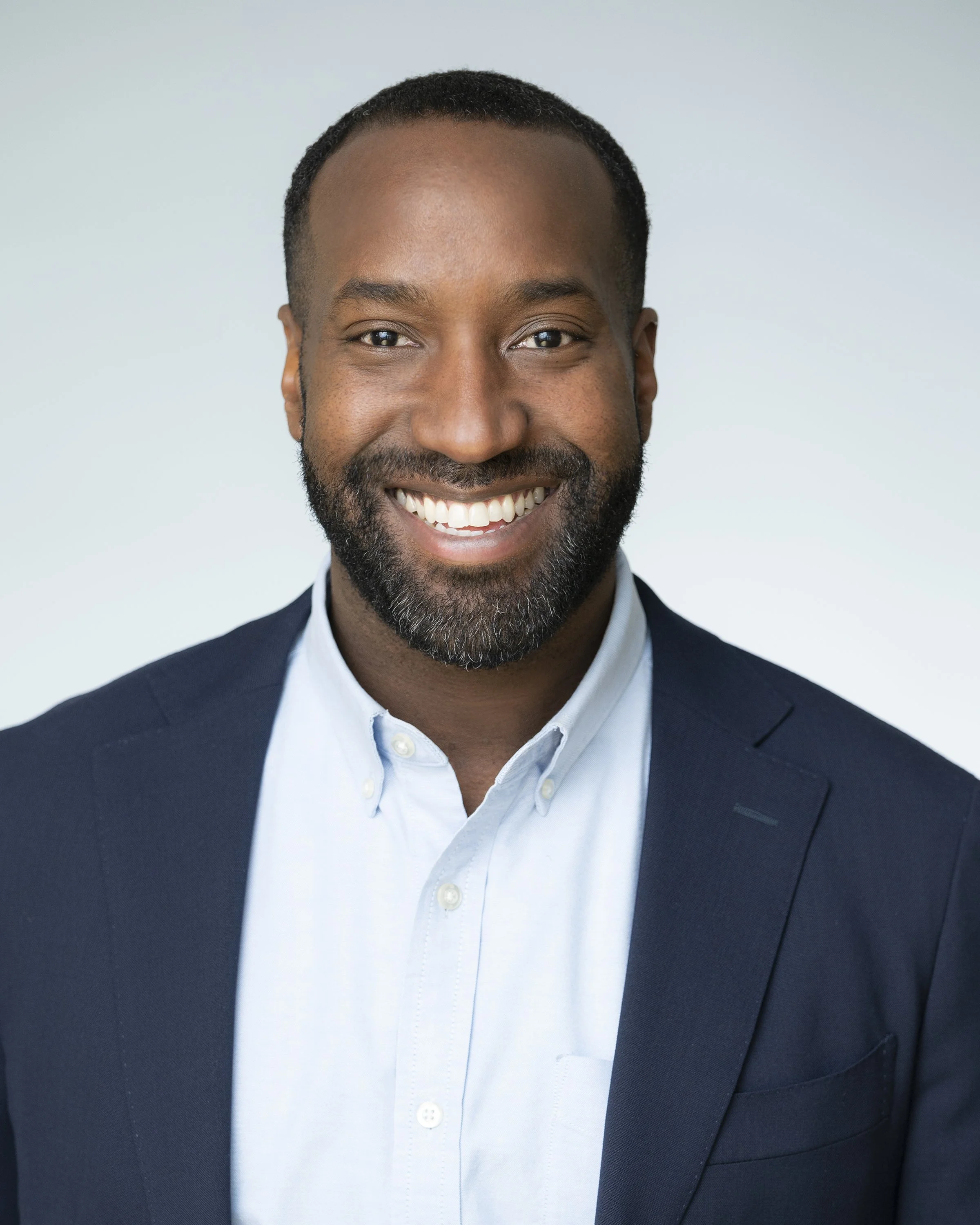 A smiling Black man with short hair and a beard wearing a light blue button-up shirt and a dark blazer, facing the camera against a plain light background.