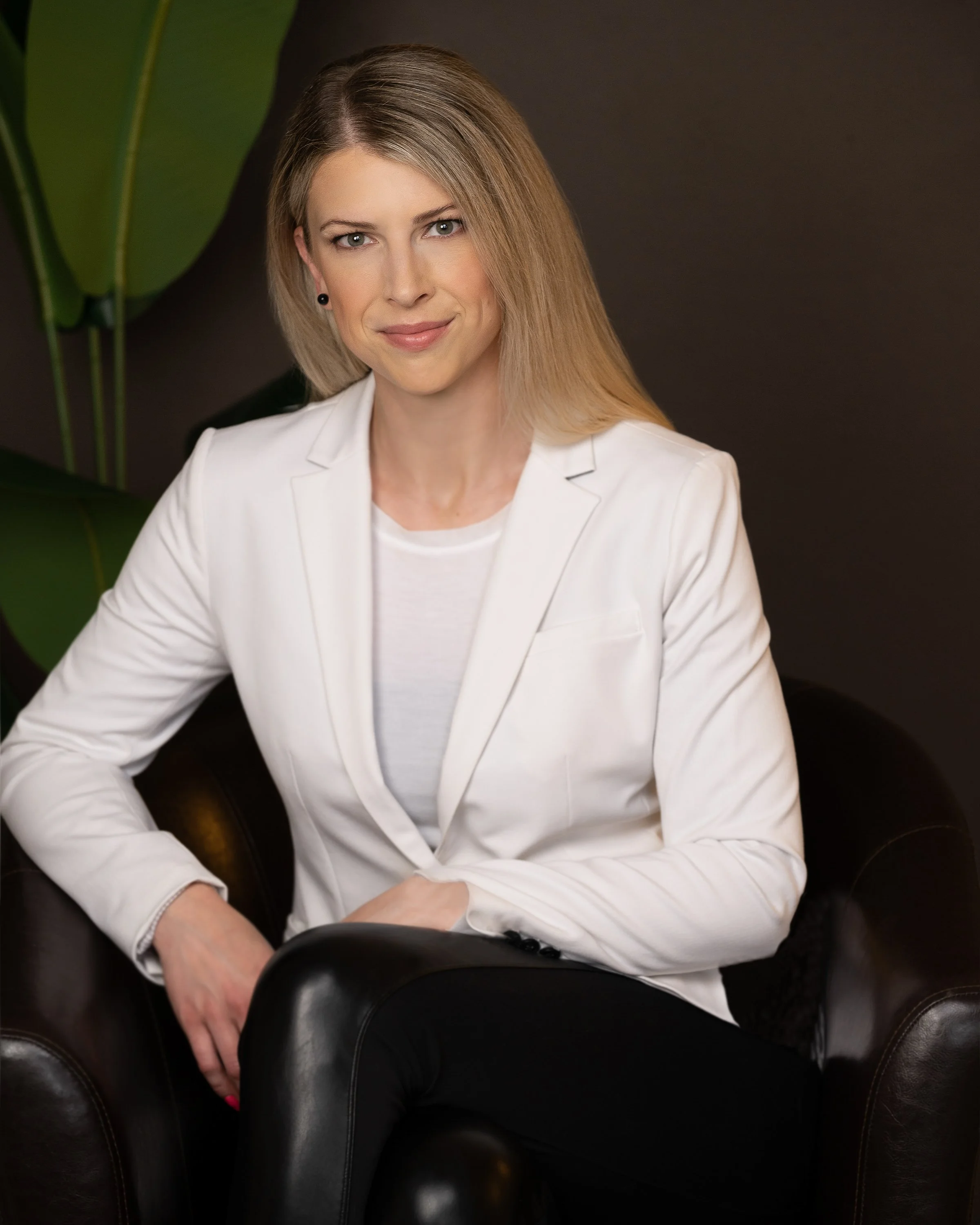 A woman with blonde hair wearing a white blazer and earrings sitting in a black leather chair with large green leaves in the background.