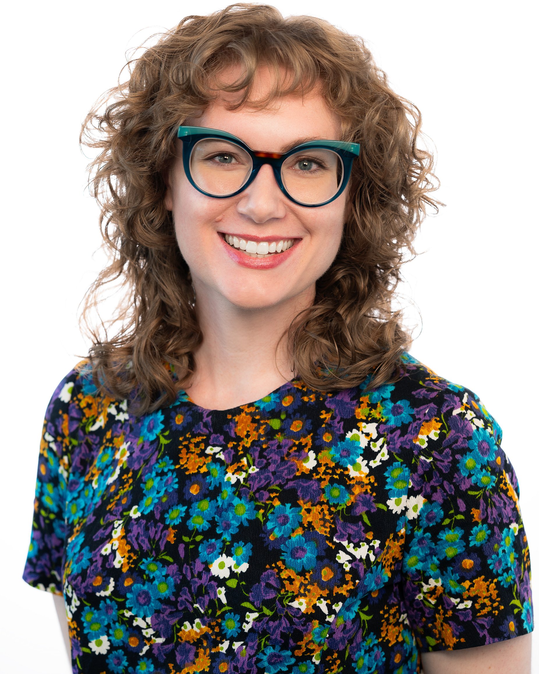 A woman with curly, light brown hair and teal-rimmed glasses smiling, wearing a multicolored floral dress with a black background.