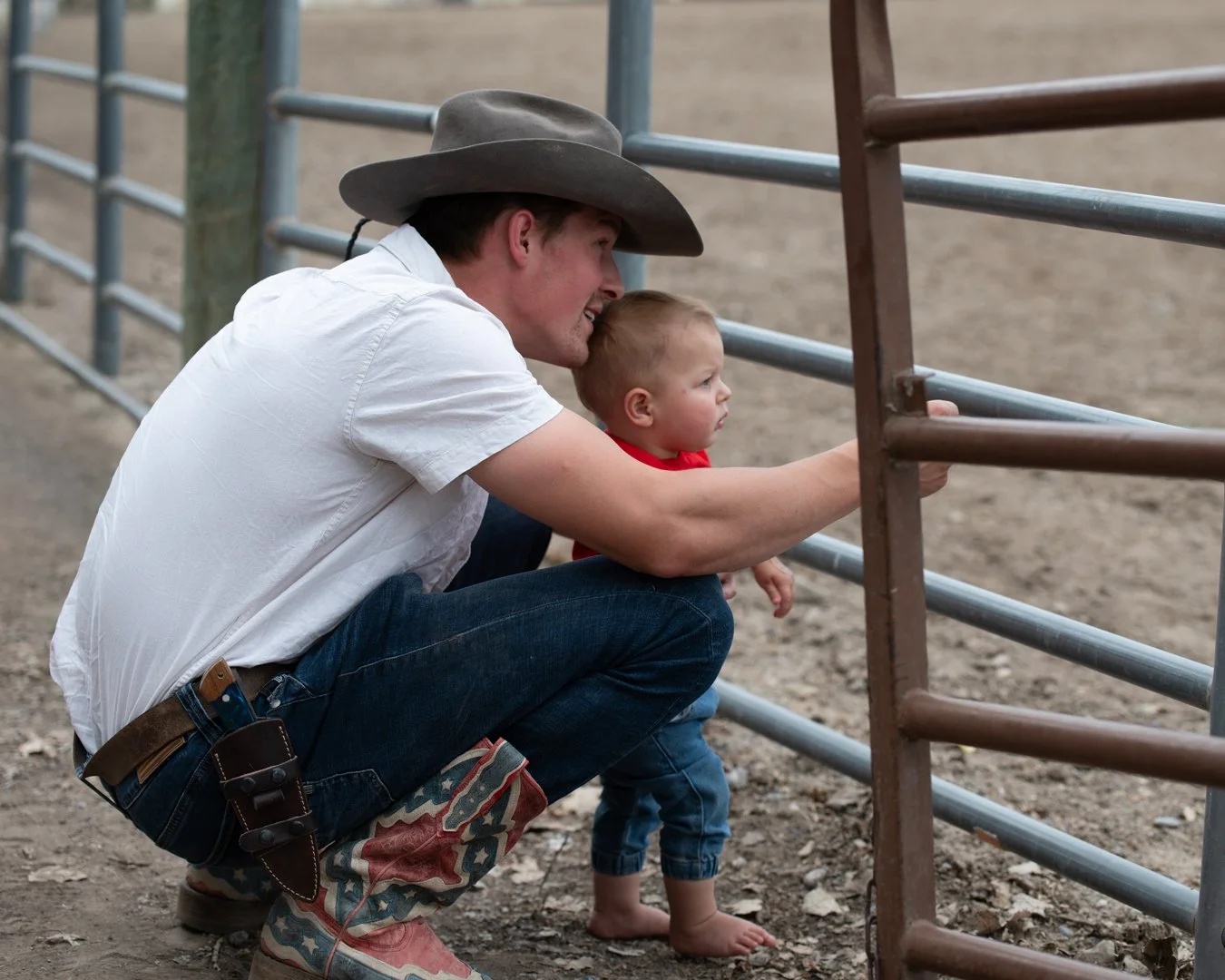 A man wearing a cowboy hat crouches next to a young boy, watching through a metal fence at a rodeo event.