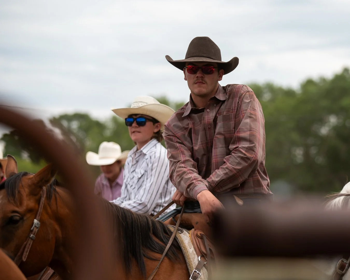 People riding horses outdoors, wearing cowboy hats and sunglasses, with a backdrop of trees and a cloudy sky.