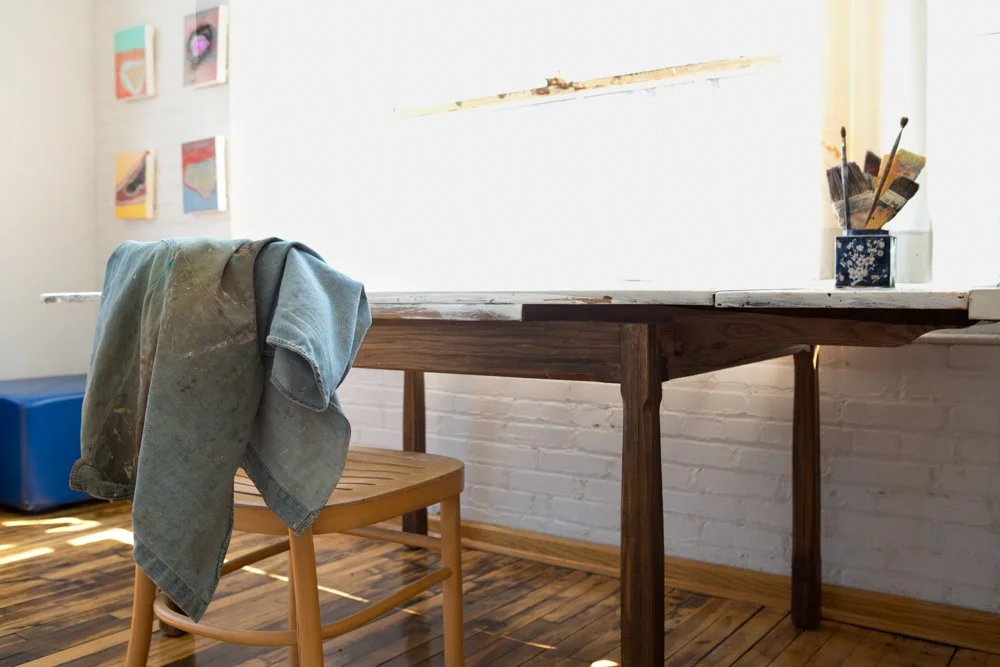 An artist's studio with a wooden table, a chair with a painter's smock draped over it, and artwork hanging on the white brick wall, with paintbrushes in a jar on the table.