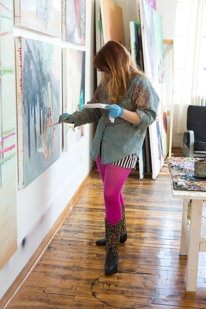A woman wearing pink tights, leopard print boots, and a paint-splattered jacket, standing and working on a painting in an art studio.