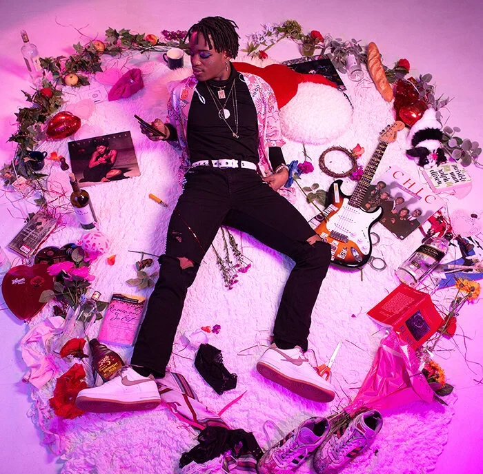 Person lying on a round white rug surrounded by various items including a guitar, flowers, books, drinks, clothes, and accessories in a colorful room with pink lighting.