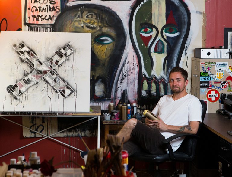 An artist sitting in his studio surrounded by colorful artwork, including a large abstract skull painting and a mixed media piece with an X pattern. The studio is decorated with stickers and paint supplies.