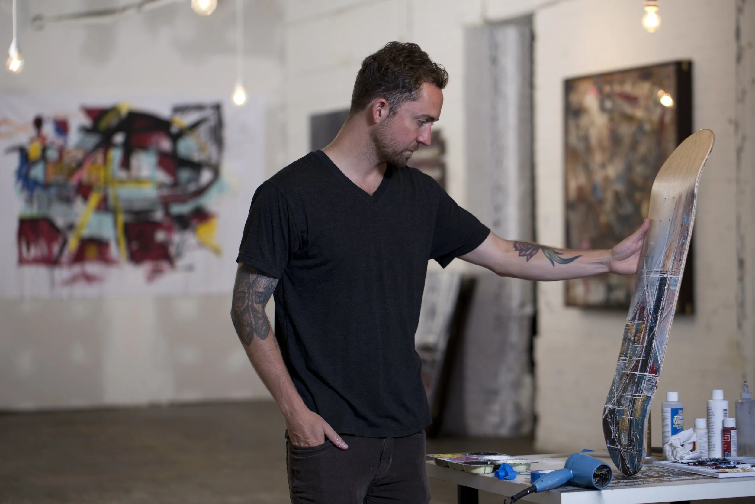 A man with tattoos on his left arm wearing a black t-shirt is looking at his art on an easel at an art studio. There are paint bottles on a table in front of him, and abstract paintings hanging on the walls.